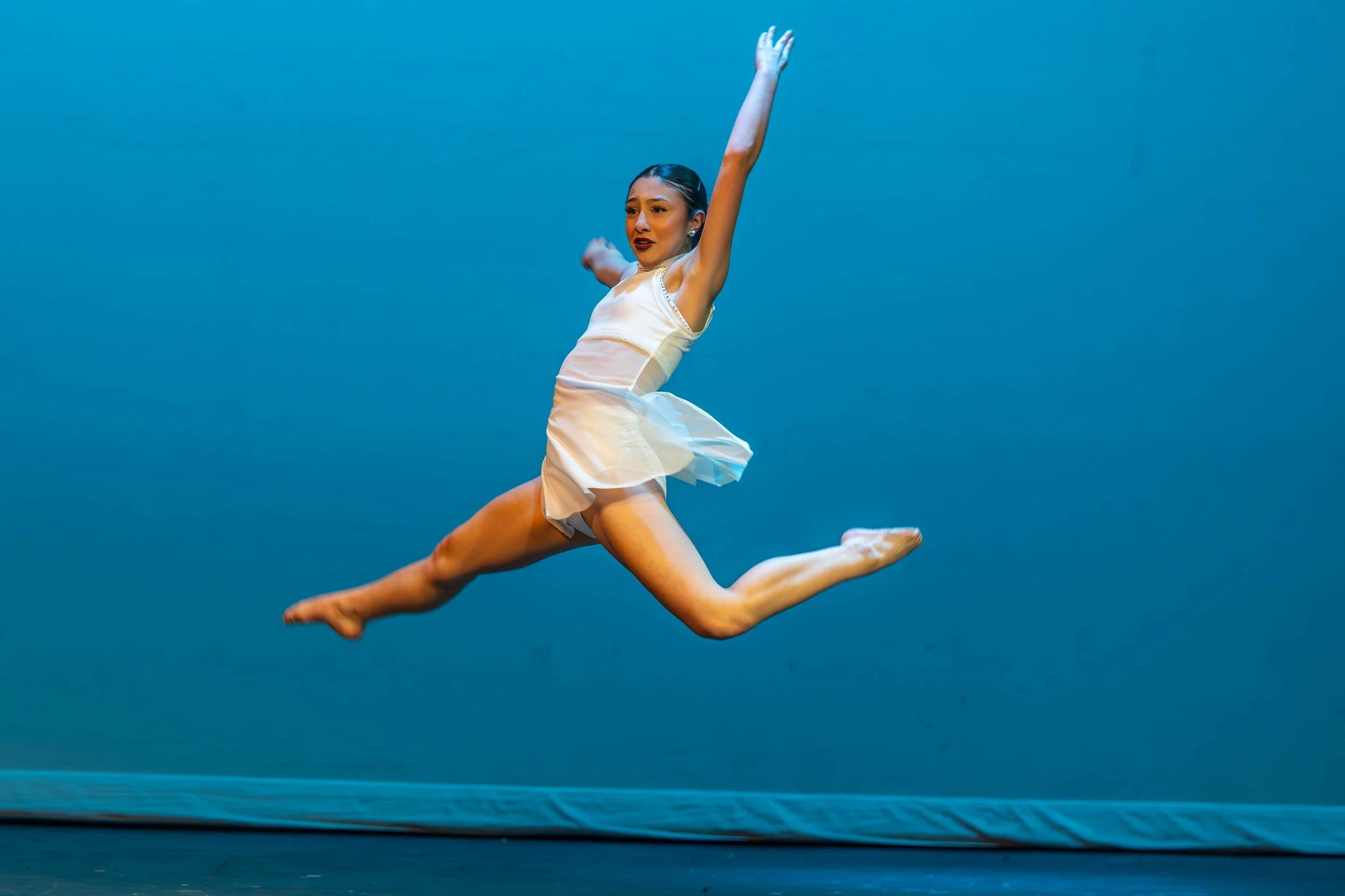 A female ballet dancer performing a leap on stage with a blue background.