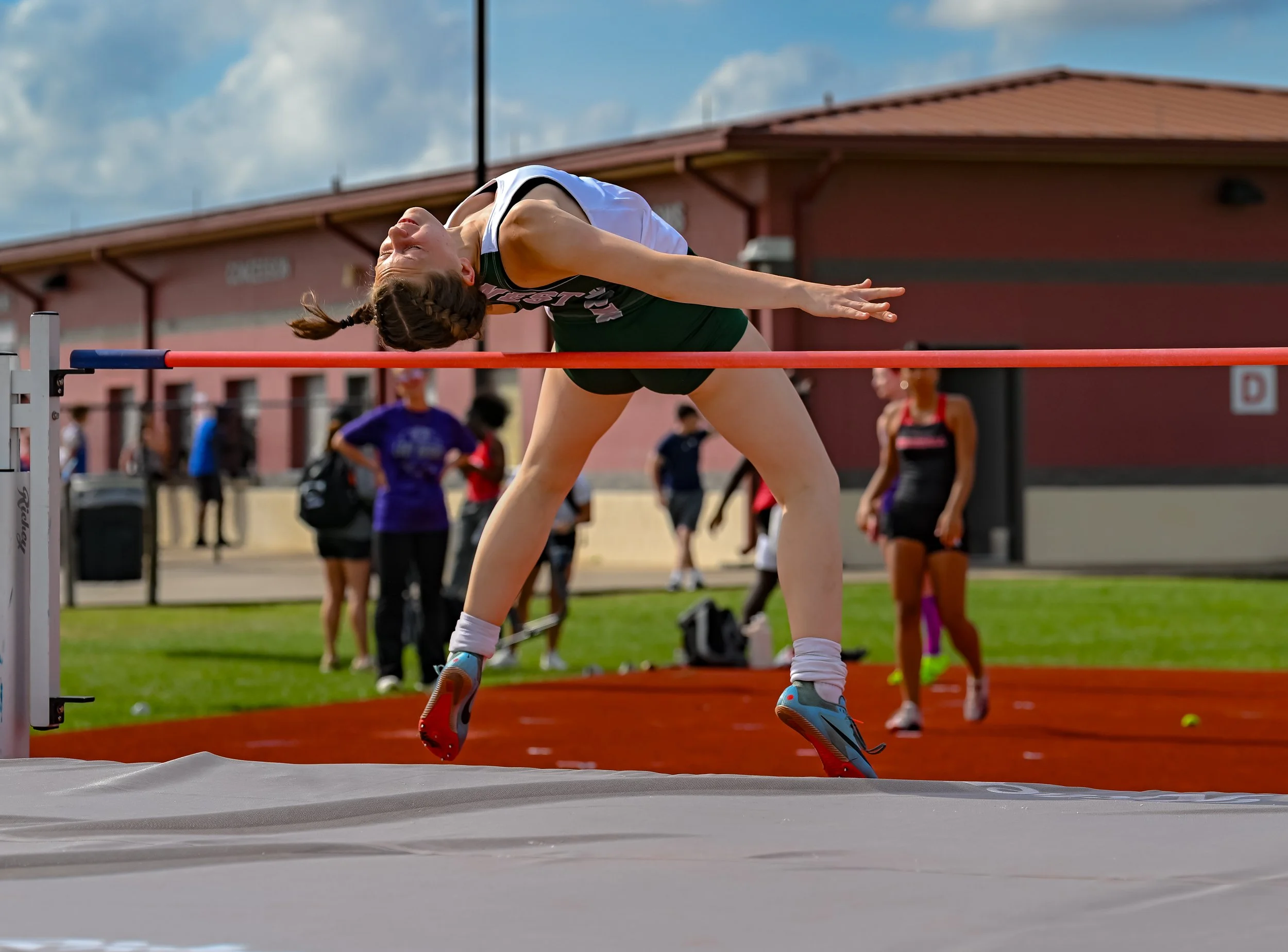 A female athlete in mid-air jumping over a hurdles during a track and field event at an outdoor stadium on a sunny day.