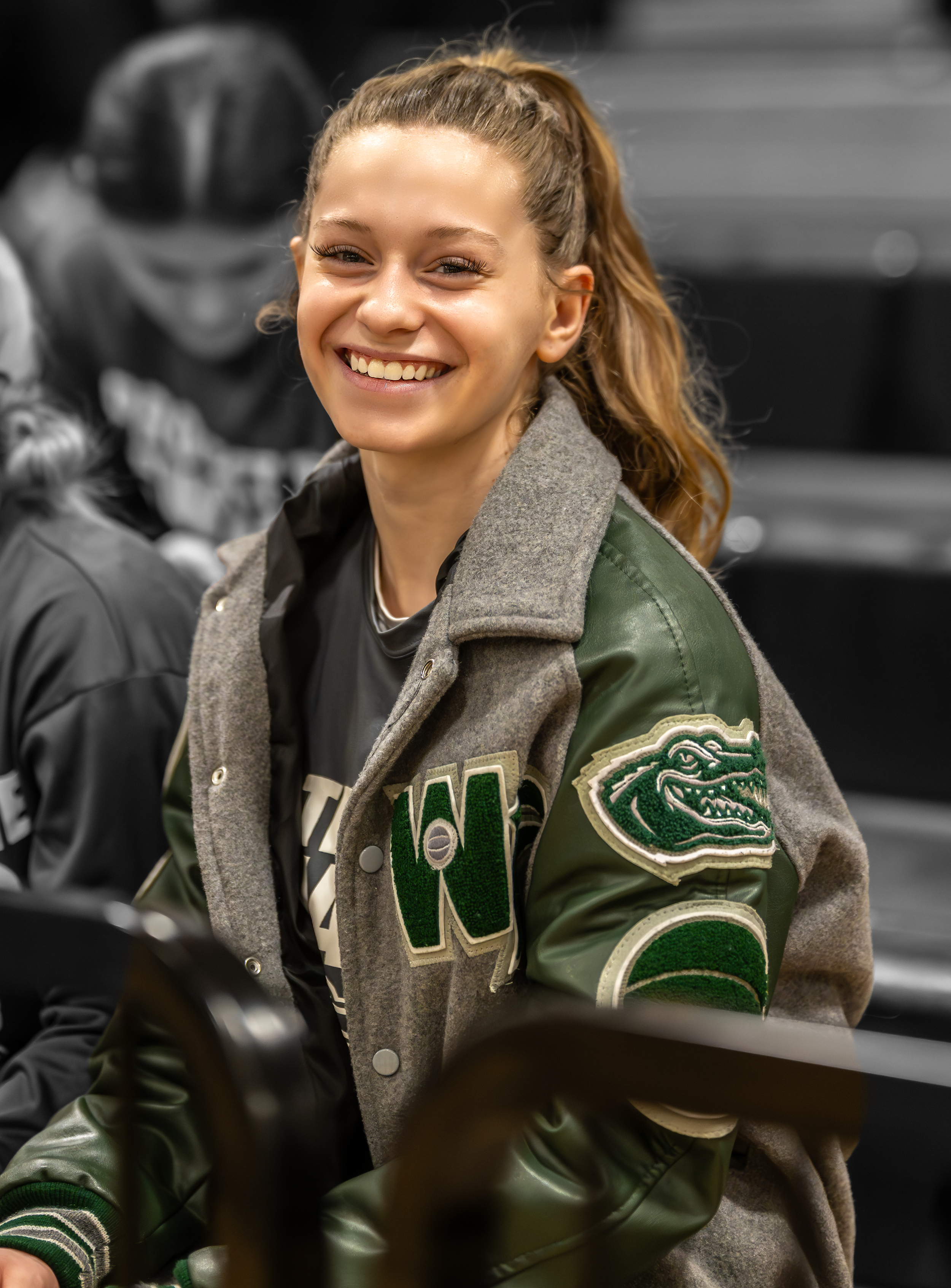 A young woman smiling with blonde hair tied back in a ponytail, wearing a green and gray varsity jacket with a dragon patch on the sleeve, sitting indoors with a blurred person and background behind her.