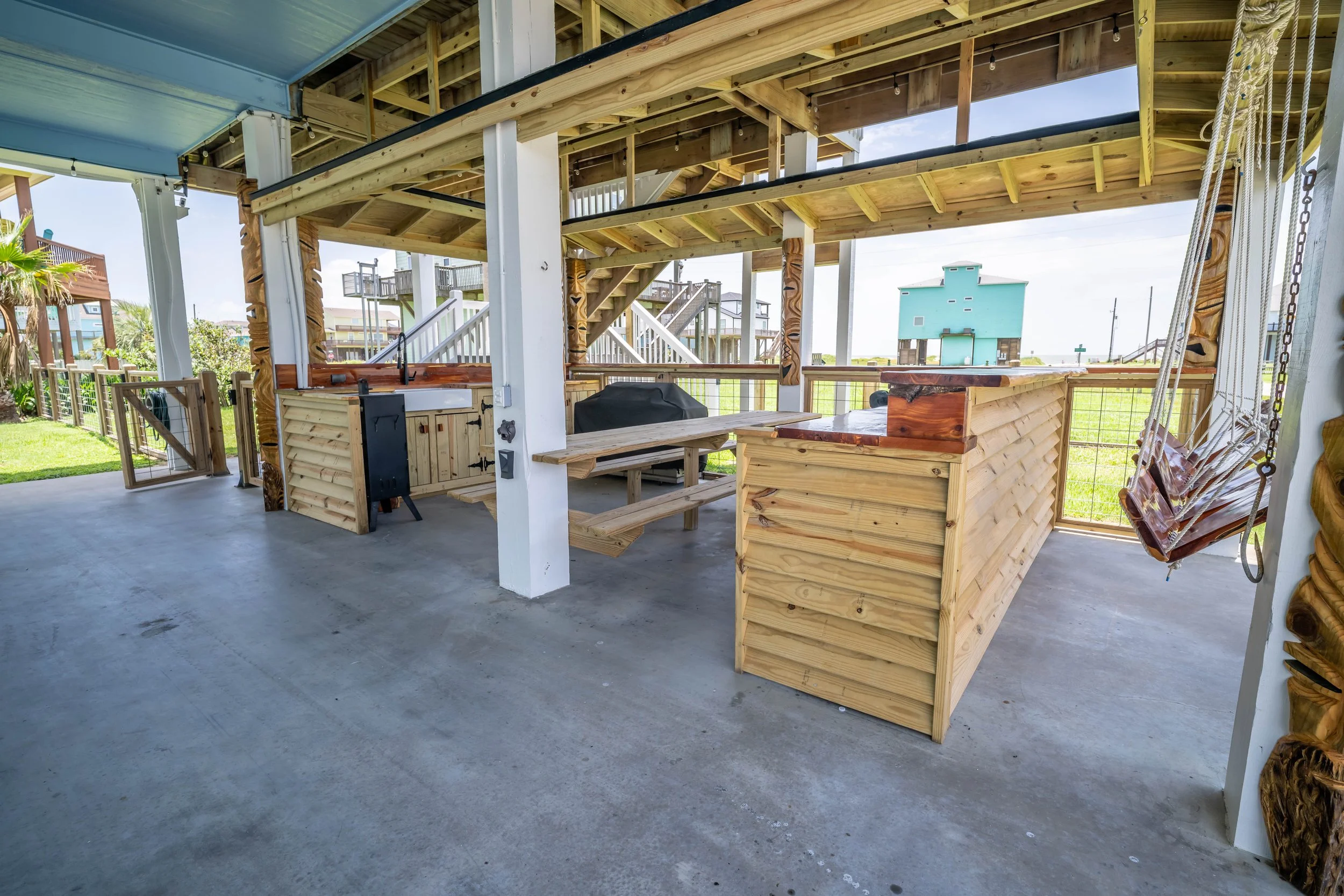 Undercover outdoor wooden deck with a built-in outdoor kitchen and seating area, large open space with concrete flooring, and houses or structures in the background.