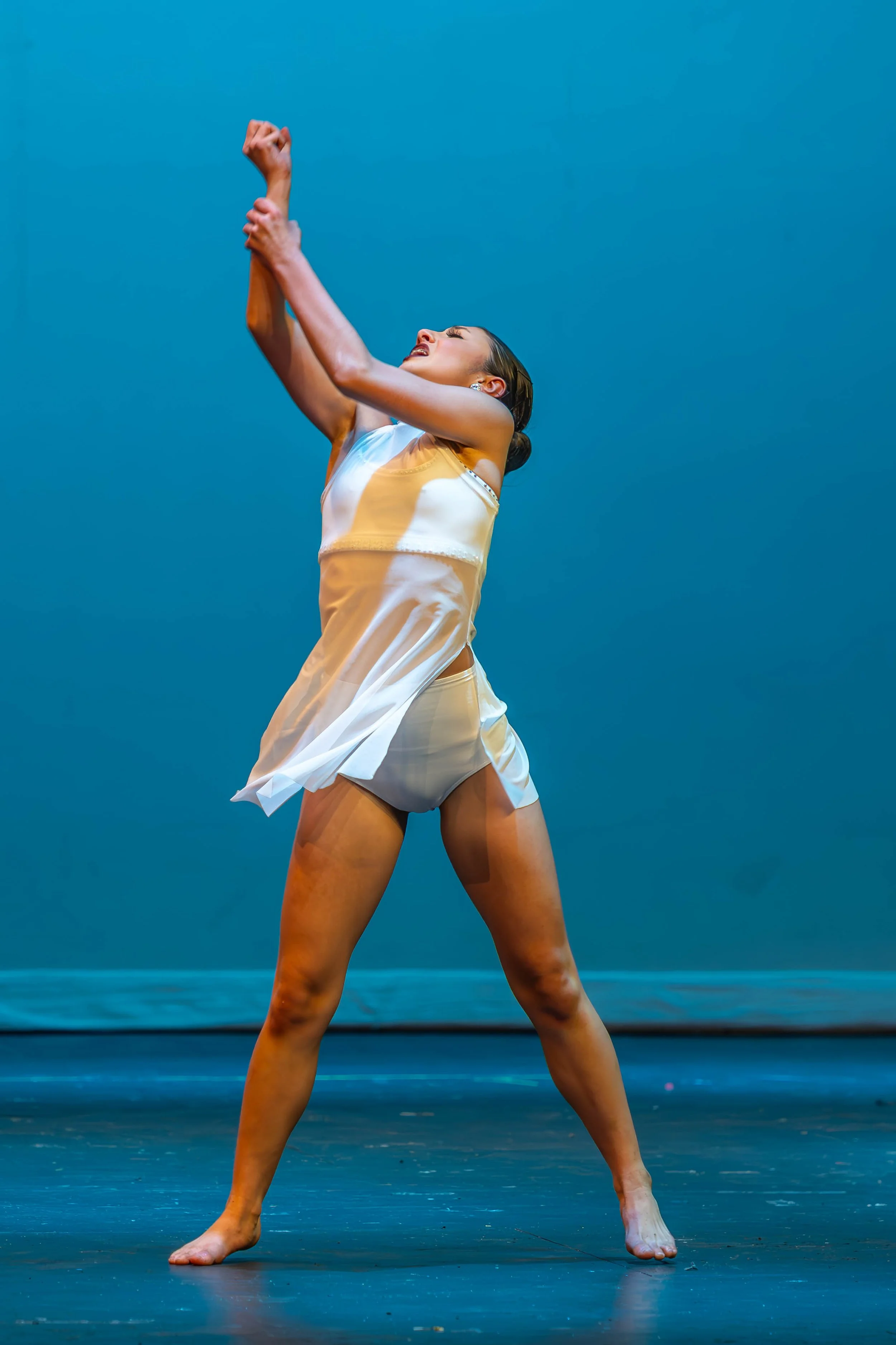 A female dancer is performing on stage against a blue background, wearing a white satin dress and striking a expressive pose with her arms raised and her body leaning back.