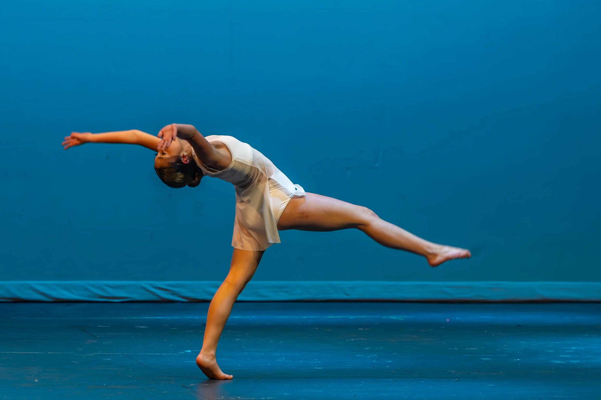 A woman in a white dress performing a contemporary dance move on a stage with a blue background, balancing on one leg with her arms extended.