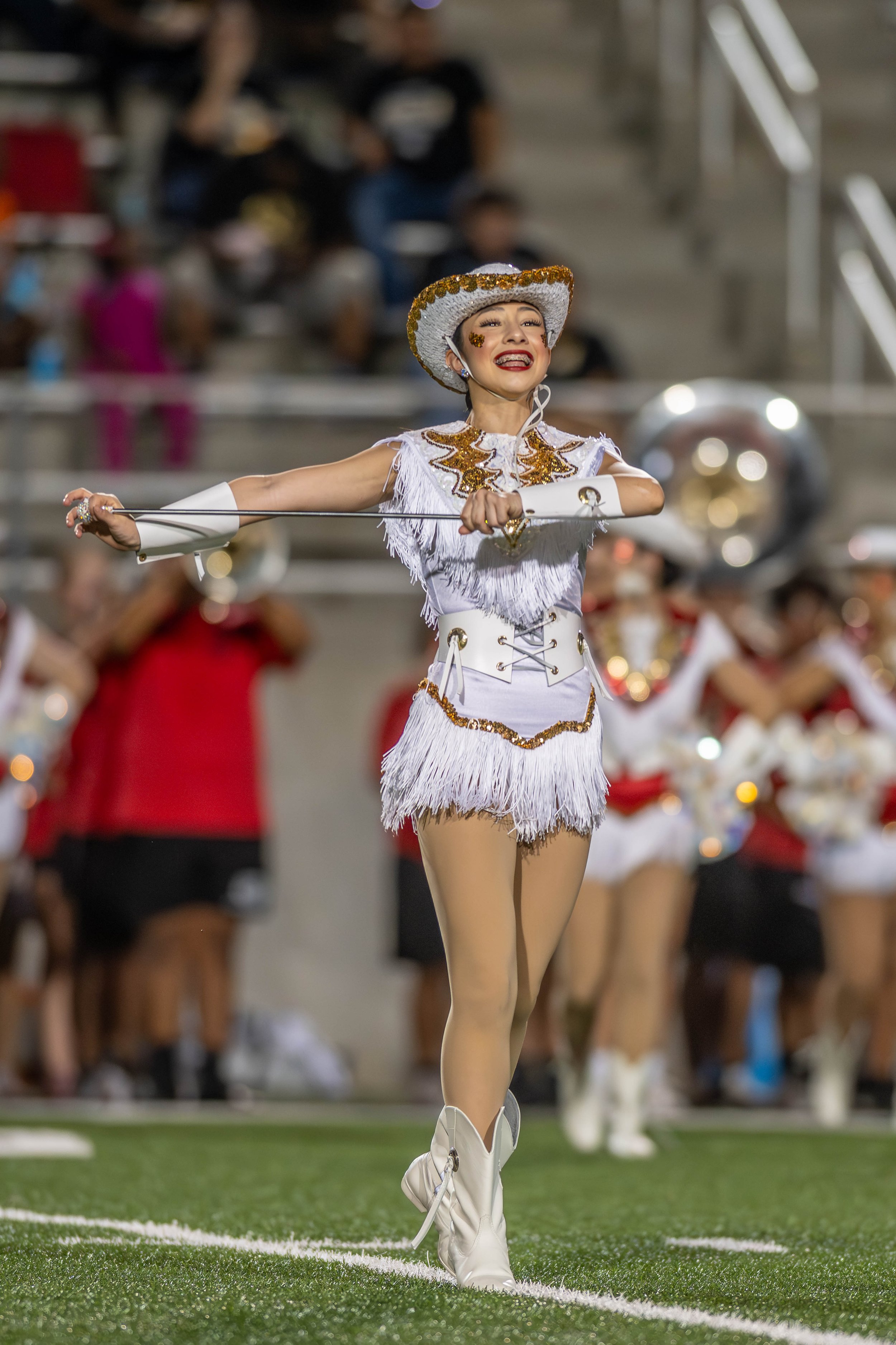 A cheerleader performing a routine at a football game, wearing a white costume with gold accents, a decorated white hat, and white cowboy boots, on a football field at night.
