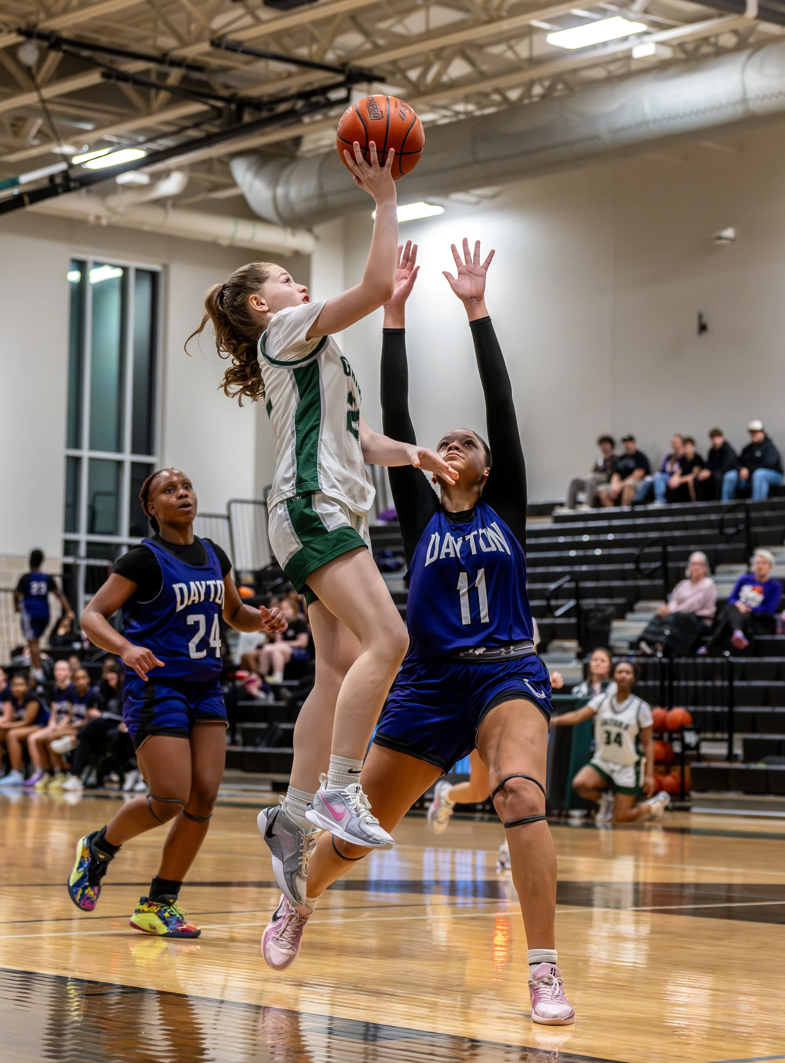 A girl in a white and green sports uniform jumping to shoot a basketball while another girl in a blue Dayton uniform defends in an indoor basketball court.