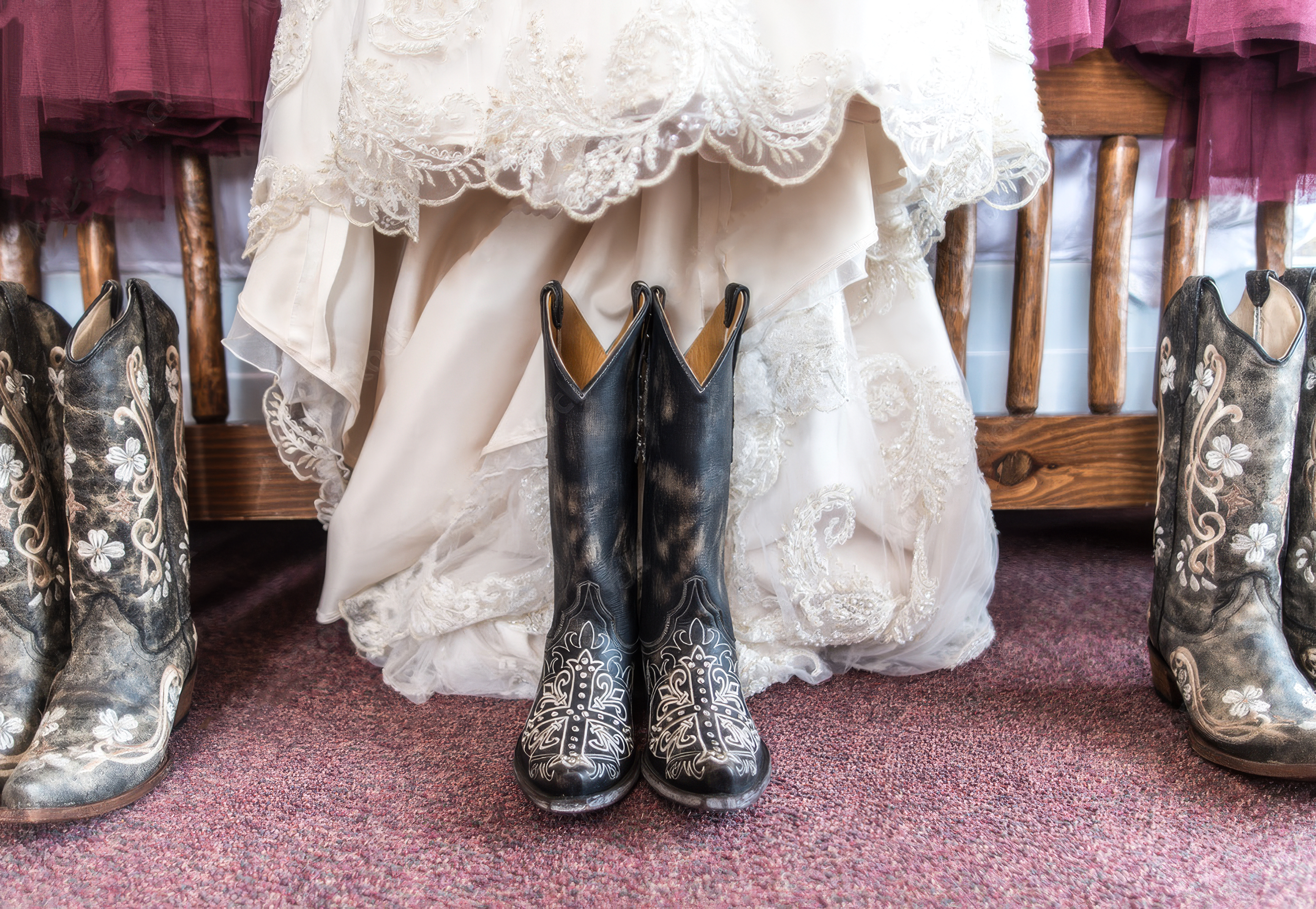A pair of black cowboy boots with white embroidered floral patterns are placed on the floor in front of a white lace wedding or special occasion dress hanging on a wooden rack. Other cowboy boots with similar embroidery are visible on either side of the main pair.