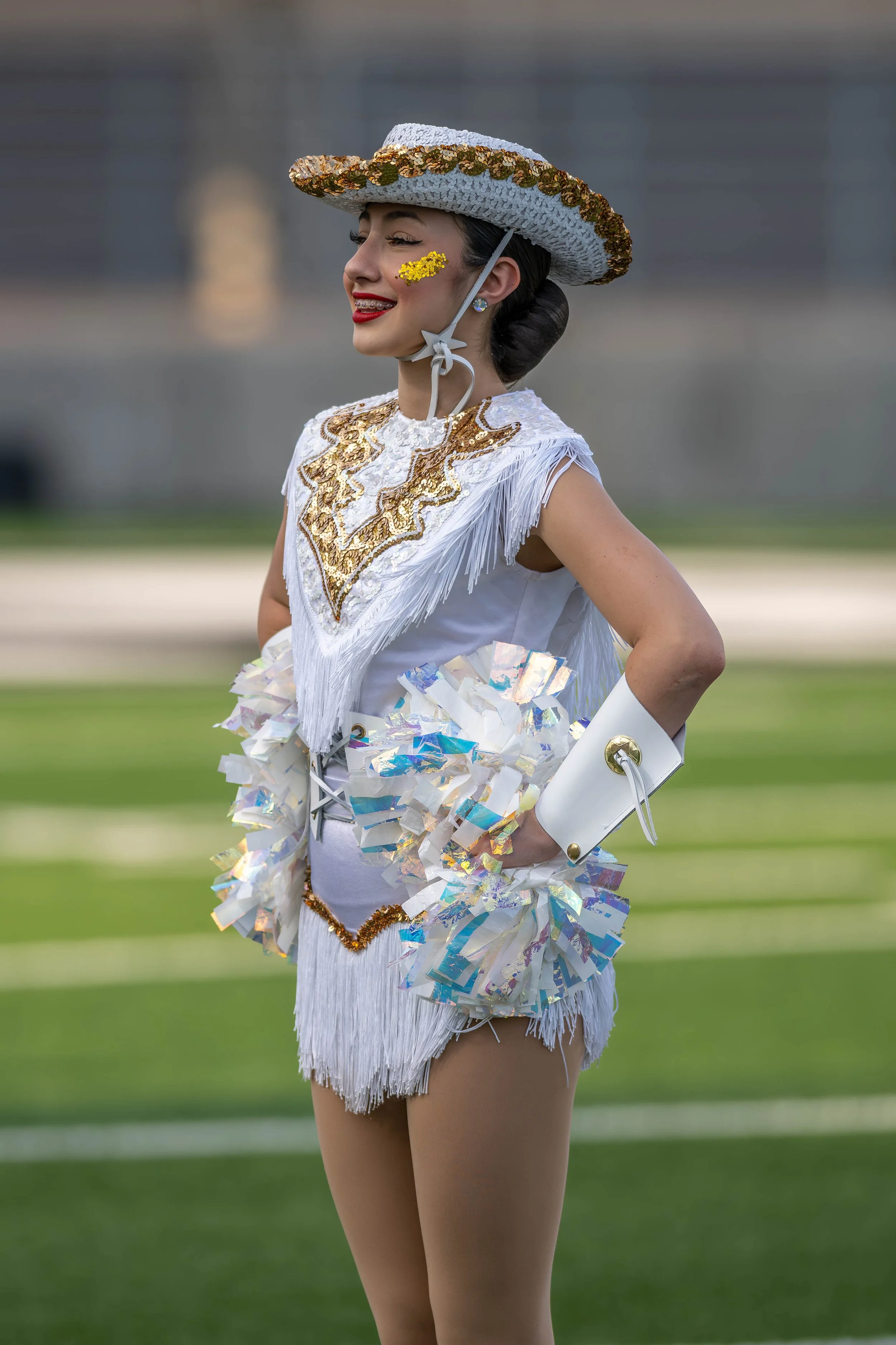 A woman in a colorful, sequined costume and a large straw hat, standing on a grassy field, with a smile and vibrant makeup.