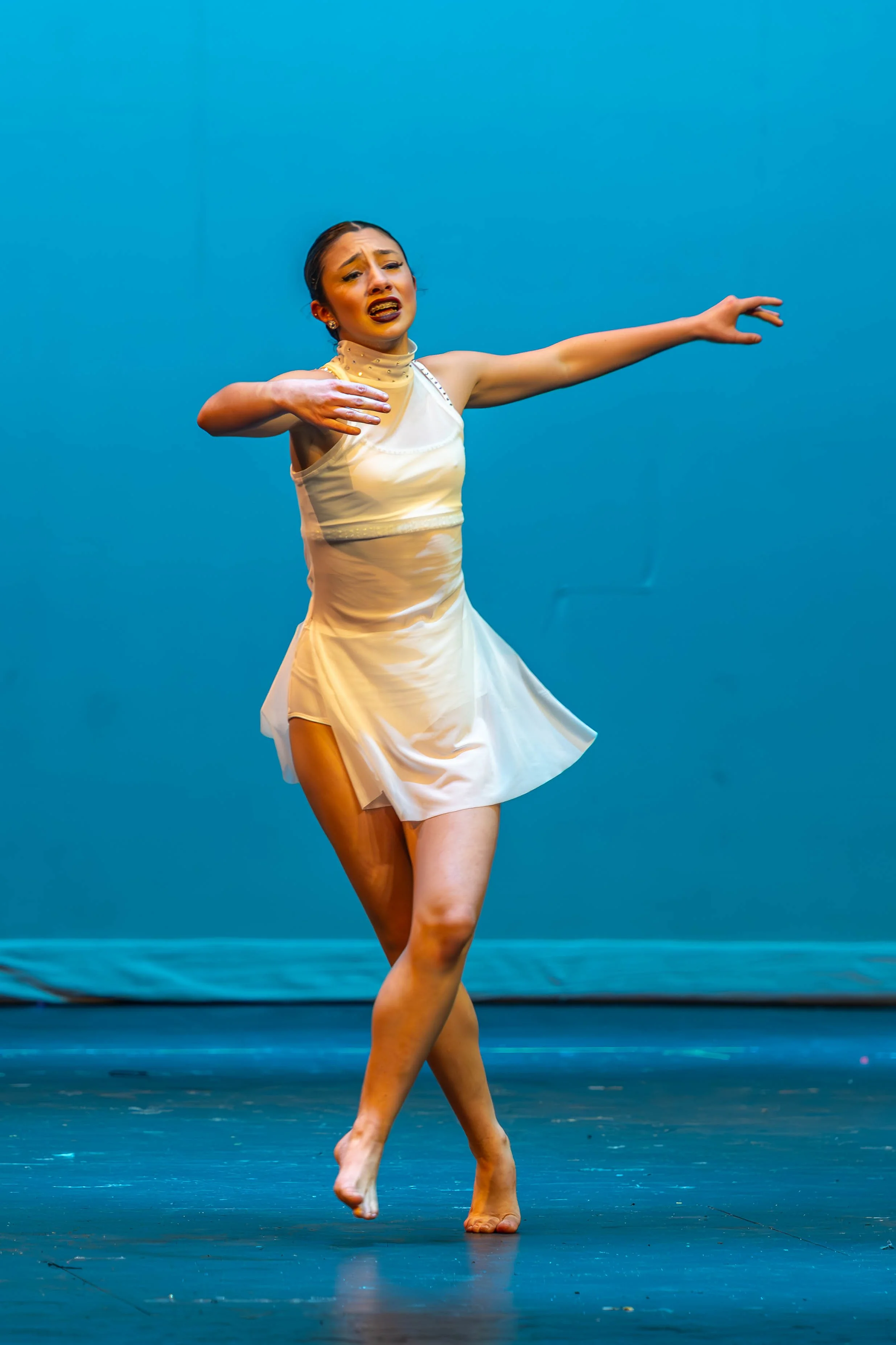 A female ballet dancer performing on stage, wearing a white dress, with an expressive face, and her arms positioned in a dance pose against a plain blue background.