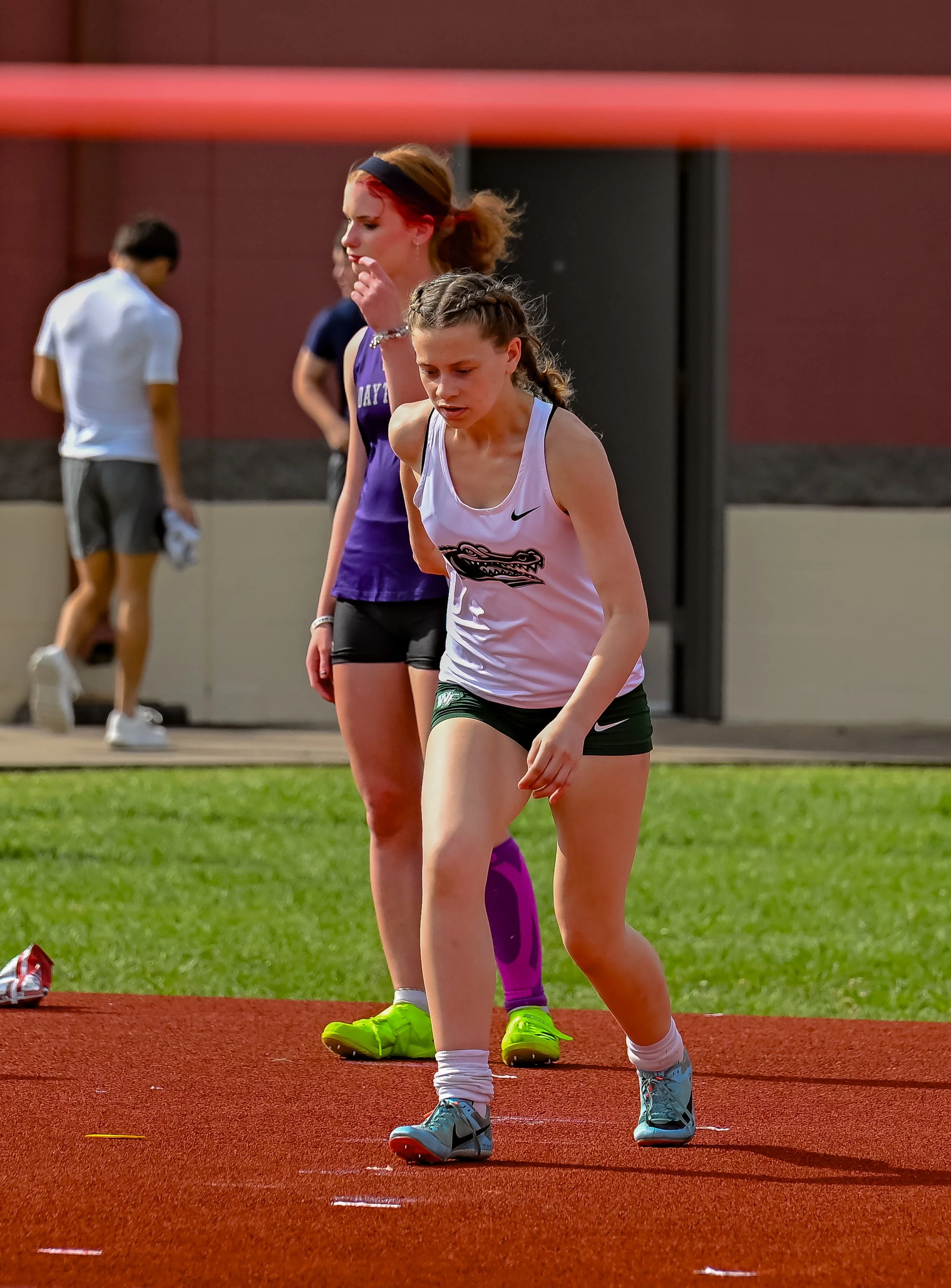 Female athletes preparing on a track field, wearing athletic outfits and shoes, with green grass and a red running track.