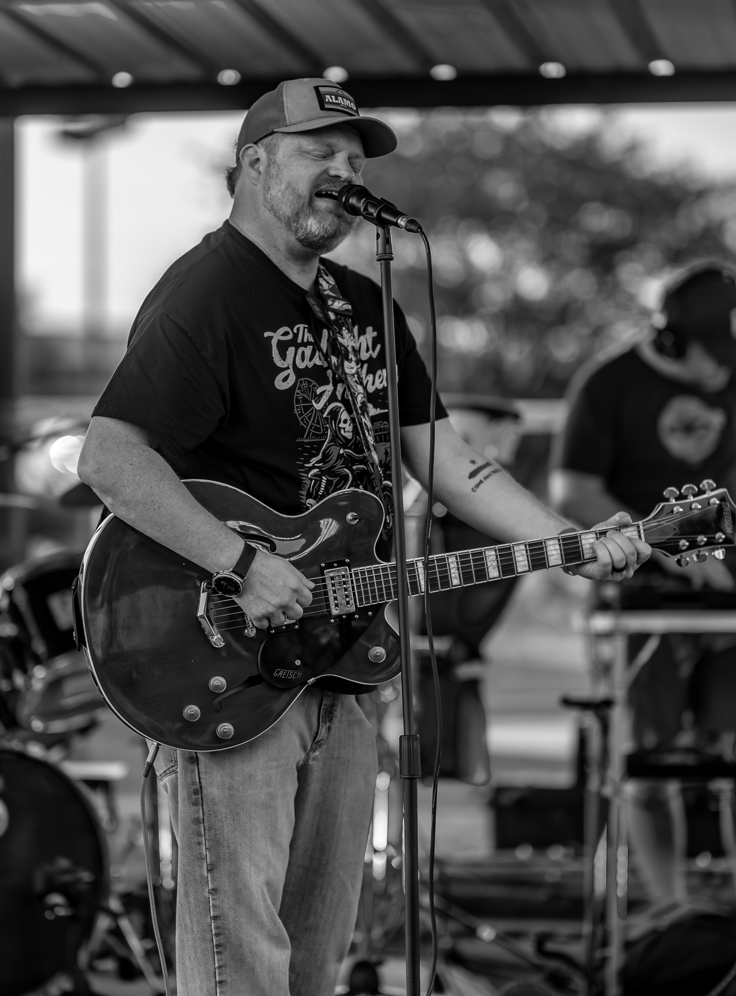 A man with a beard and mustache singing into a microphone while playing an electric guitar at an outdoor event, wearing a cap and casual clothes.
