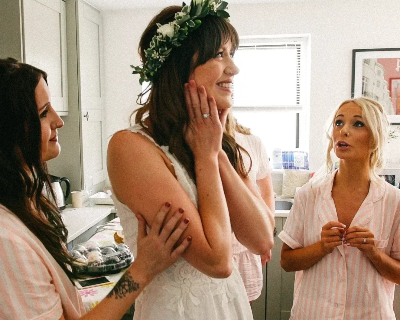 A bride with leaf and flower crown smiling while bridesmaids in pink pajamas surround her in a kitchen, preparing for her wedding.