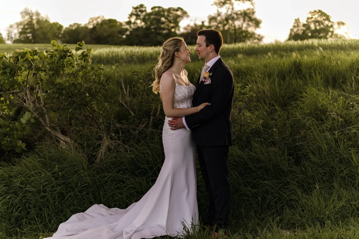 A bride and groom stand close together in a grassy field during sunset, gazing at each other, with trees in the background.