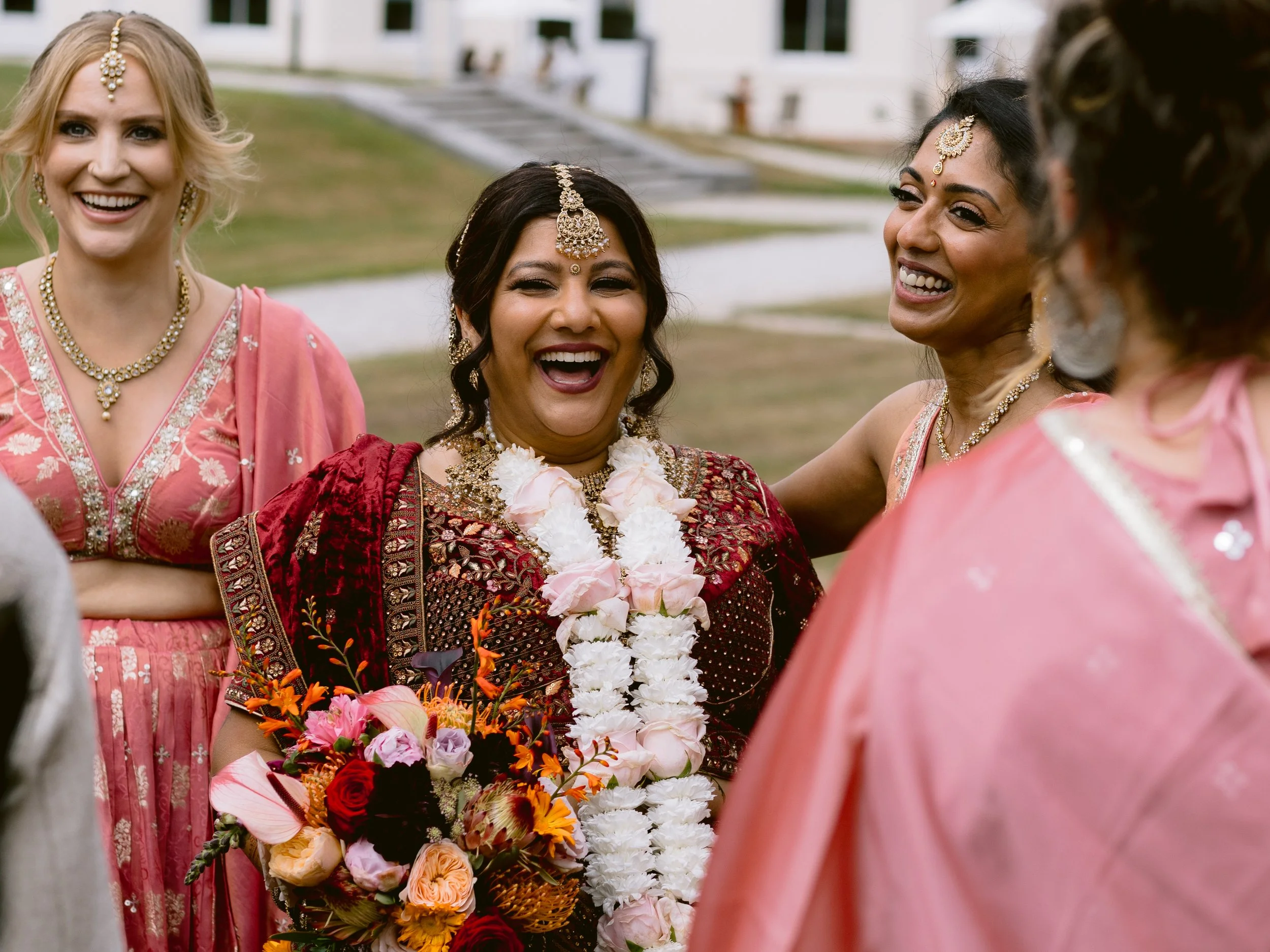 A bride in South East Asian wedding attire laughing with a group of her friends and family with Oakley hall in the background