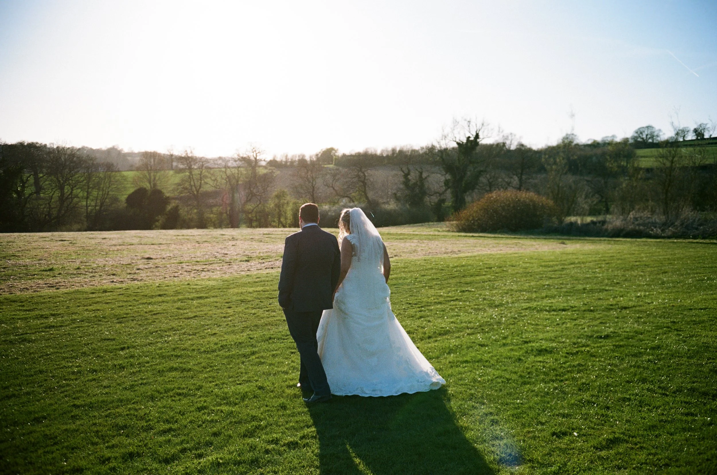 A bride and groom walking hand in hand across a grassy field with trees and hills in the background, during daylight hours.