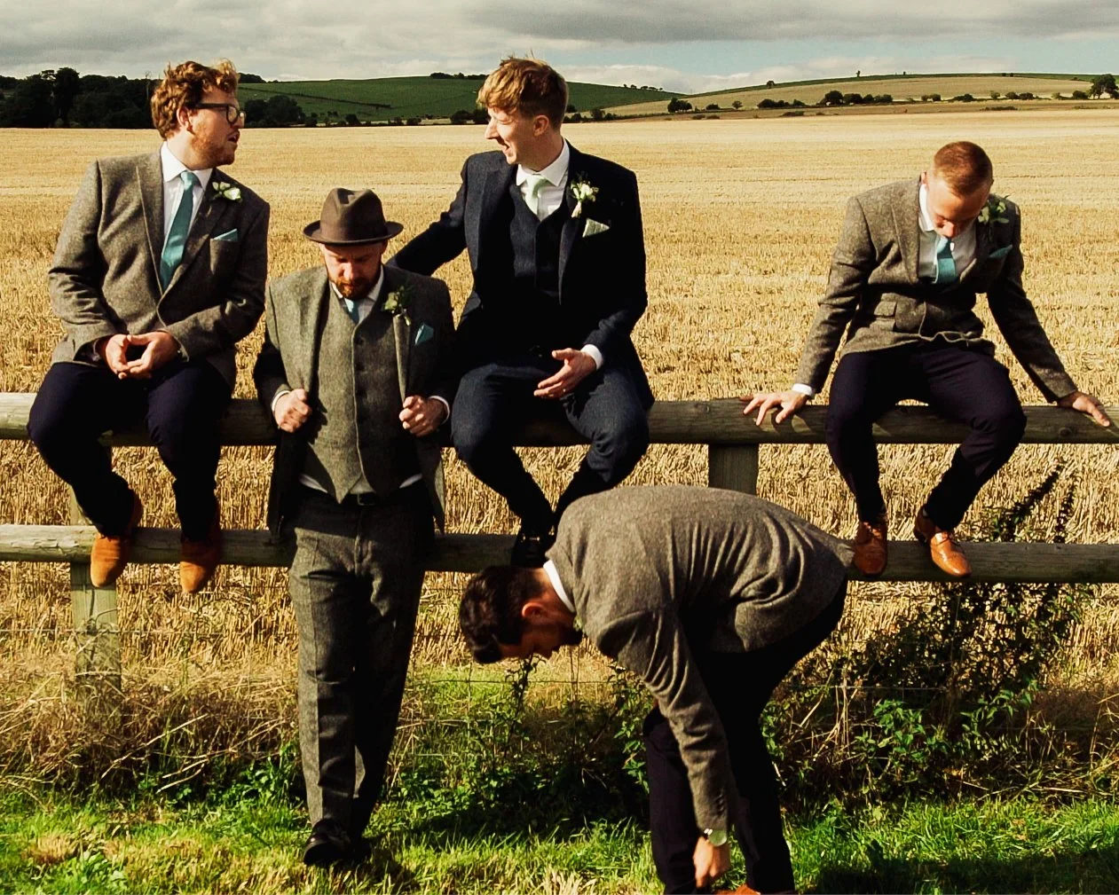 A candid image of groomsmen in suits posing on a wooden fence against rolling green countryside.
