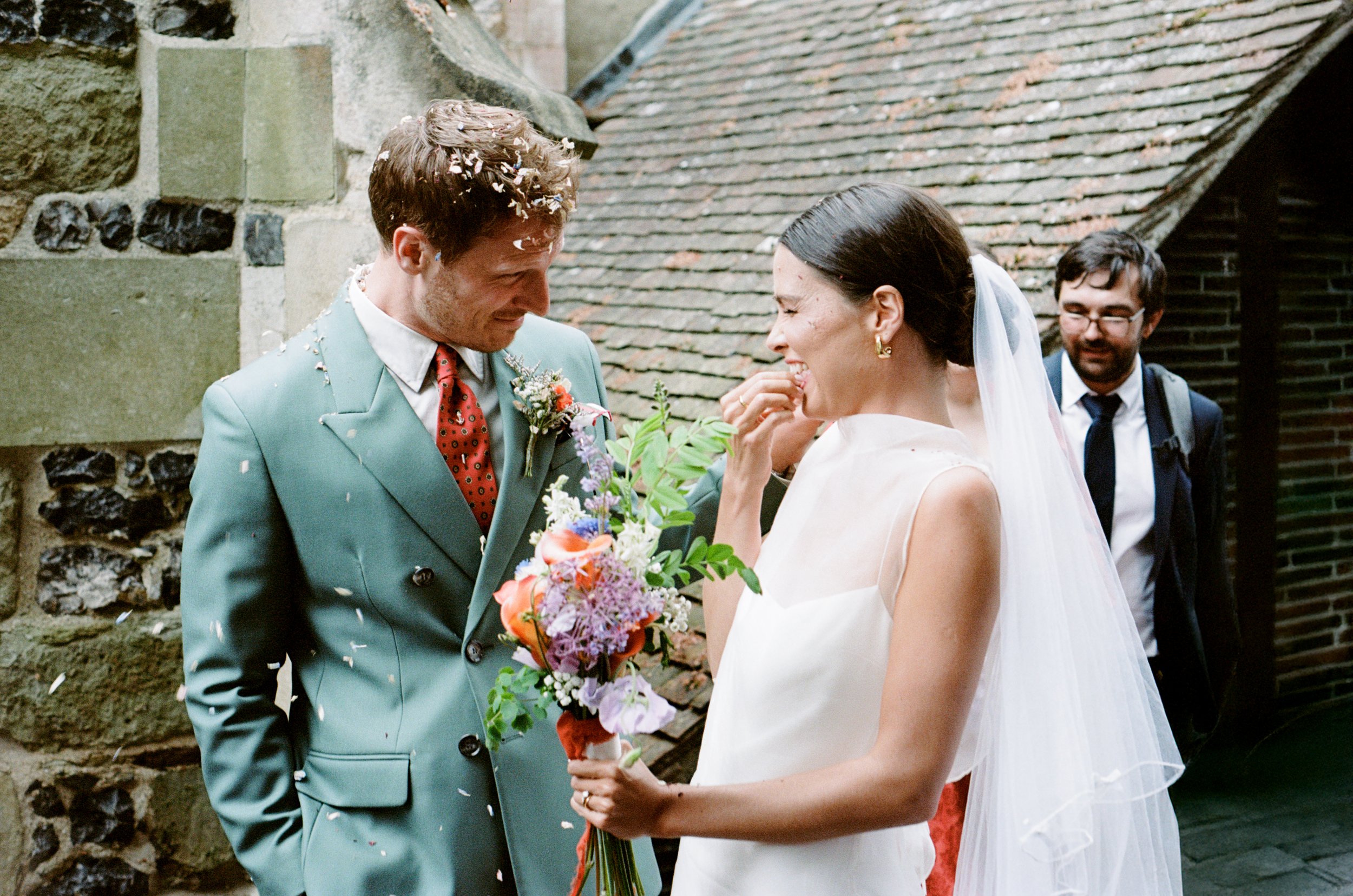 A bride and groom sharing a moment during their wedding, with a man in the background, while flowers and confetti are present.