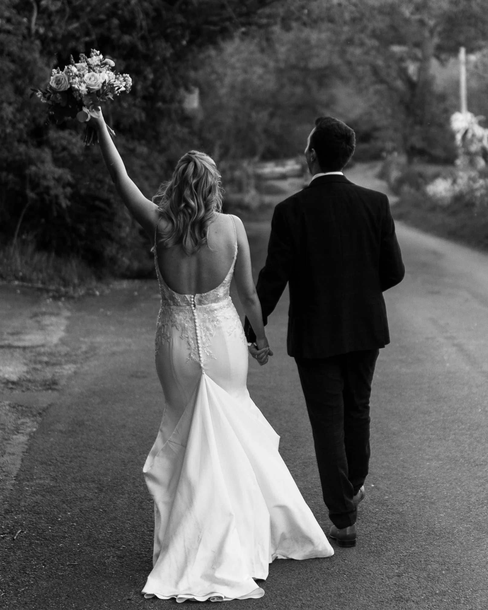 A black and white image of a groom and bride walking away and the bride's hand is carrying a bouquet of flowers