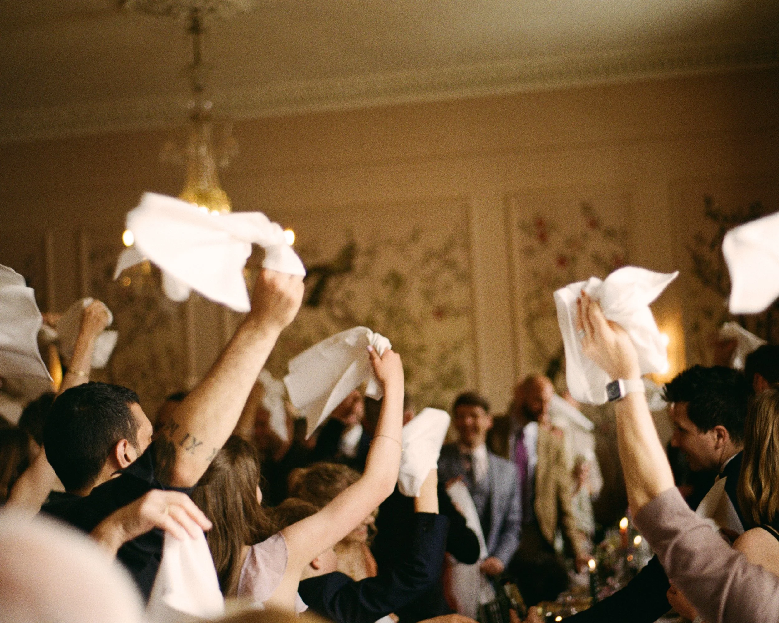Wedding guests waving white napkins beneath a chandelier during a lively evening reception.