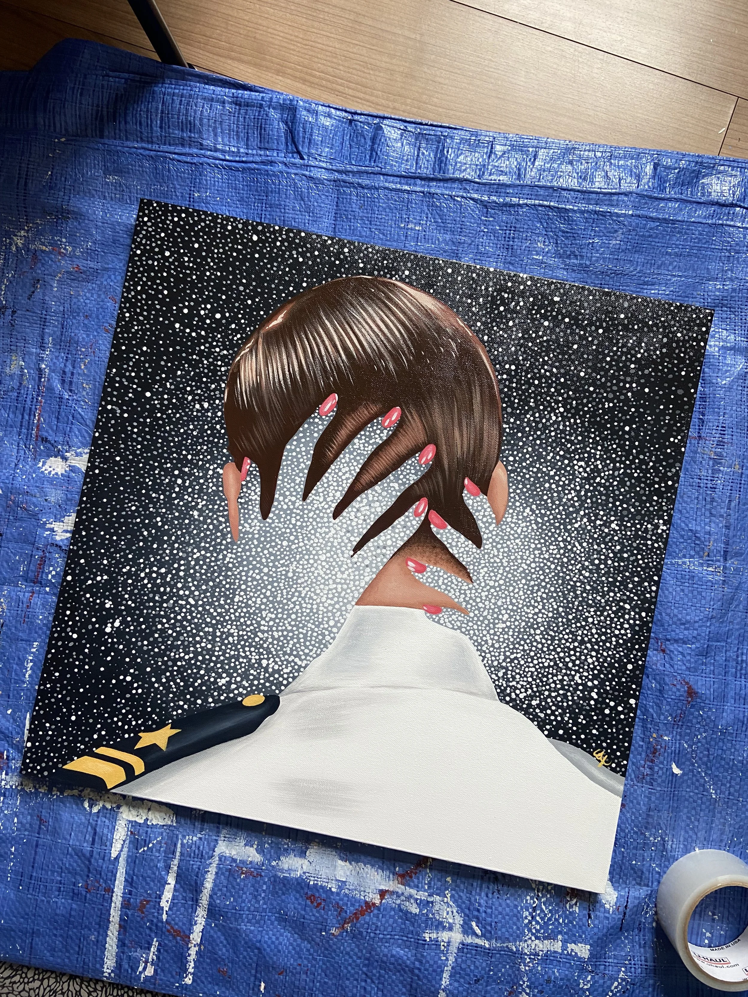 A colorful artwork on a canvas features a woman with brown hair, holding her hand to her face, set against a starry black background, with her nails painted pink and her hand adorned with rings. The canvas is placed on a blue tarp, and a roll of tape