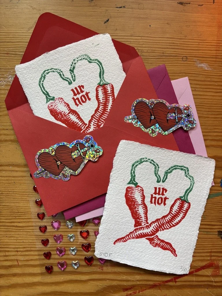 Valentine cards with chili pepper designs and the phrase 'ur hot,' surrounded by heart-shaped confetti, on a wooden surface.
