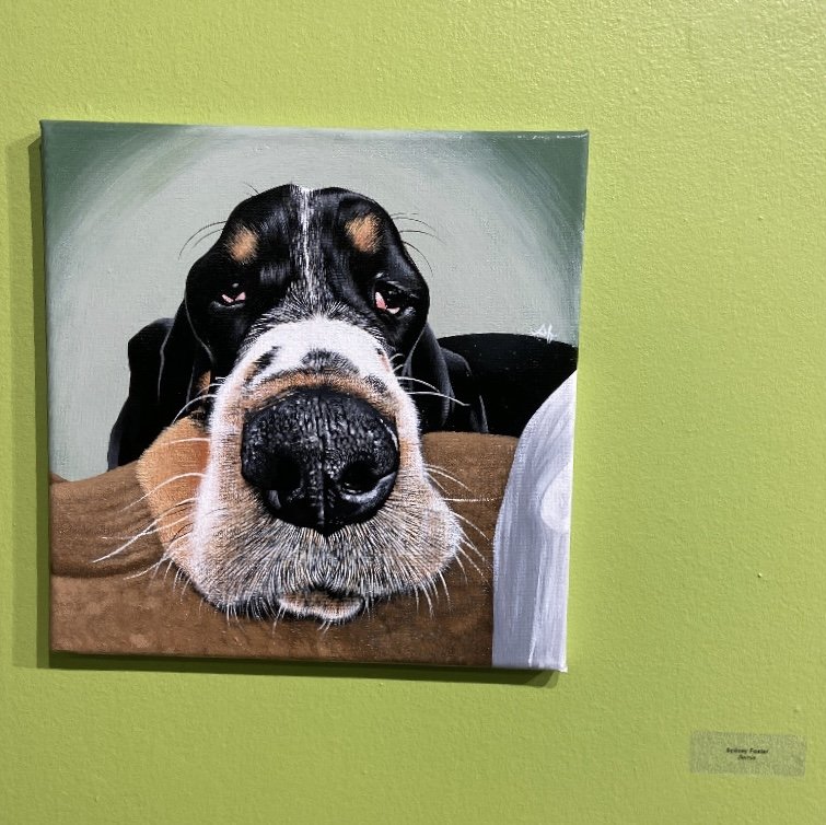 Close-up painting of a basset hound in a realistic style, resting its head on a brown surface, set against a light green wall.