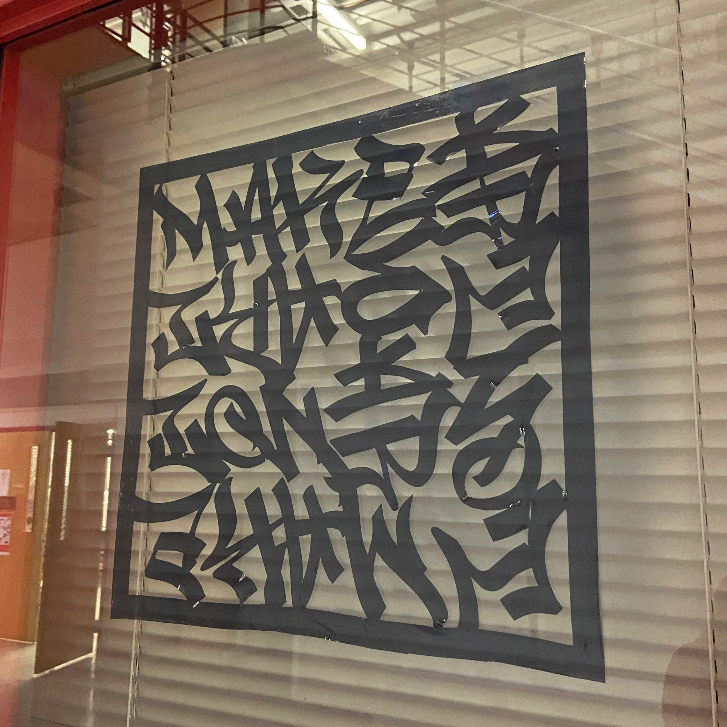 A large decorative wall hanging with black abstract Arabic calligraphy on a dark background, mounted on a wall with horizontal textured panels.