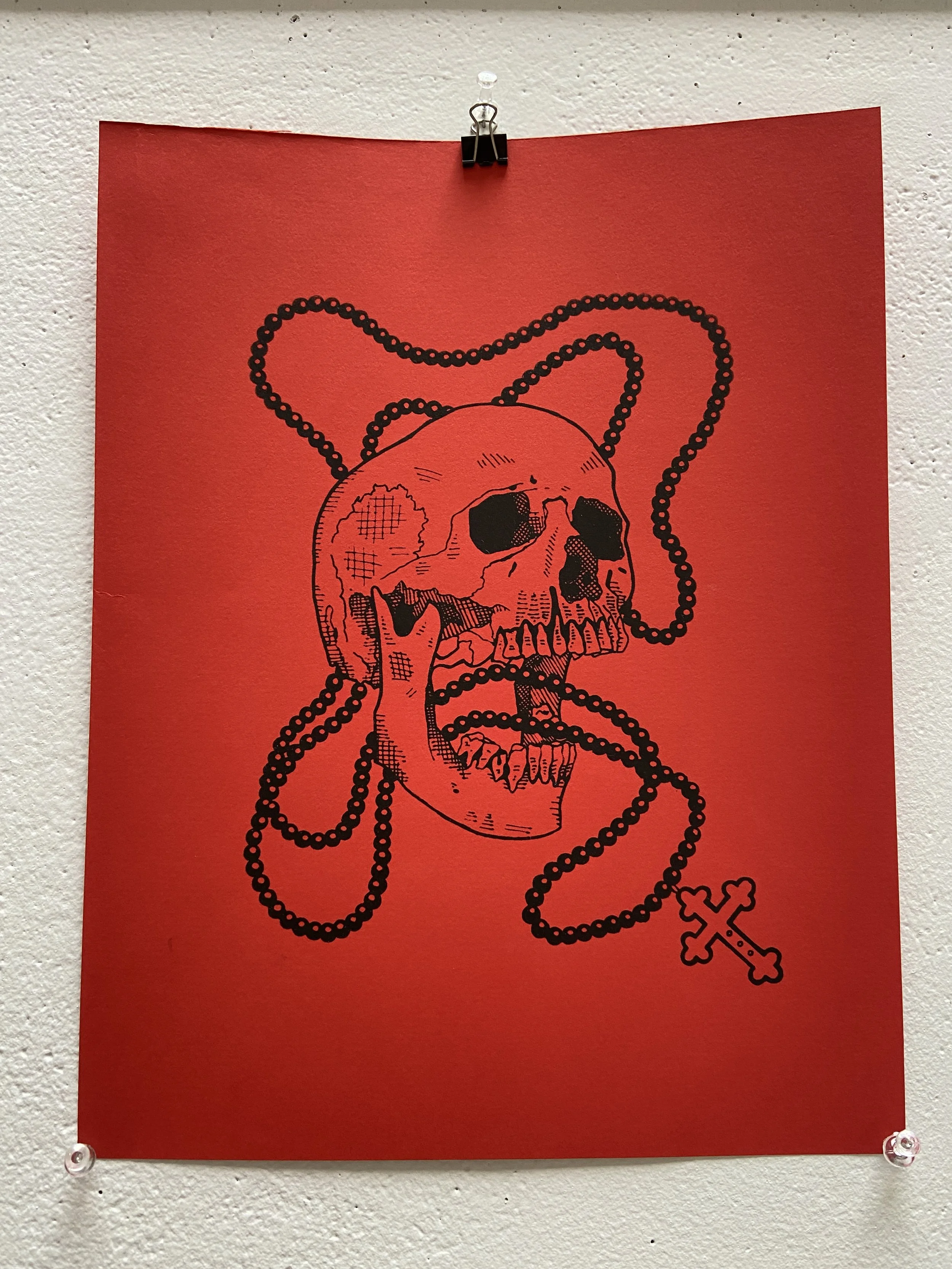 A red piece of paper with a black skull and chain design taped on a white textured wall.