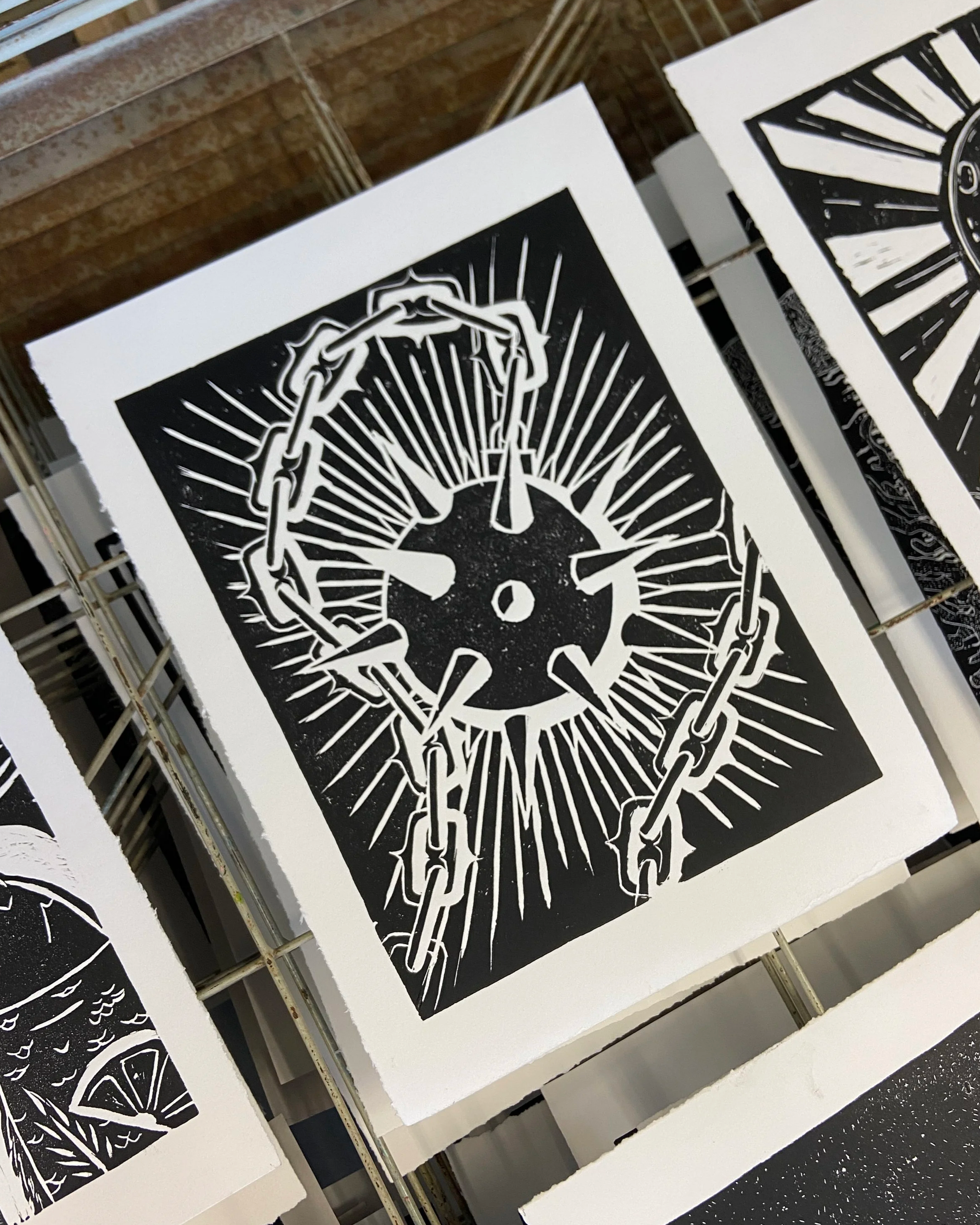 Black and white print of a broken chain surrounding a clock face with a clock hand pointing at 12:00, sun rays emanating from behind the clock.