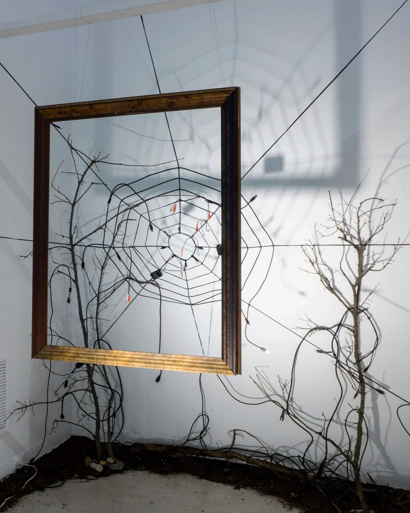 A decorative installation featuring leafless trees, LED lights, and a large spider web made of wiring within a wooden frame, set in an indoor space with white walls.