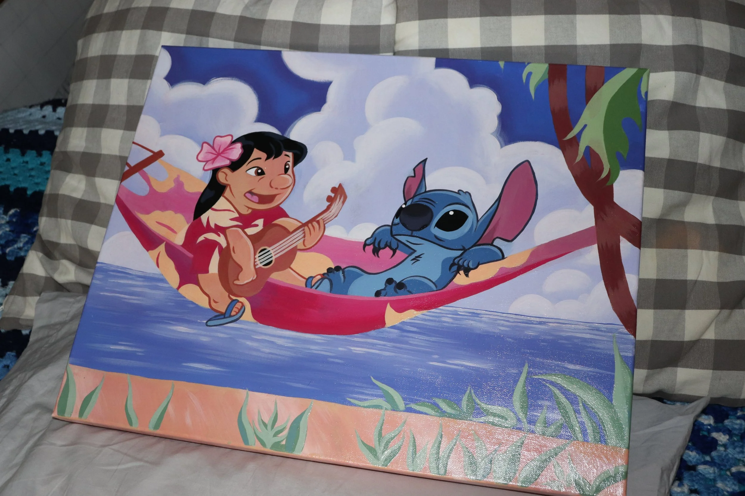 Illustration of Moana with her hair in a pink flower playing a guitar on a pink hammock. Stitch from Lilo & Stitch is lying on the hammock with big ears, surrounded by a tropical setting with rainbows, clouds, and trees.