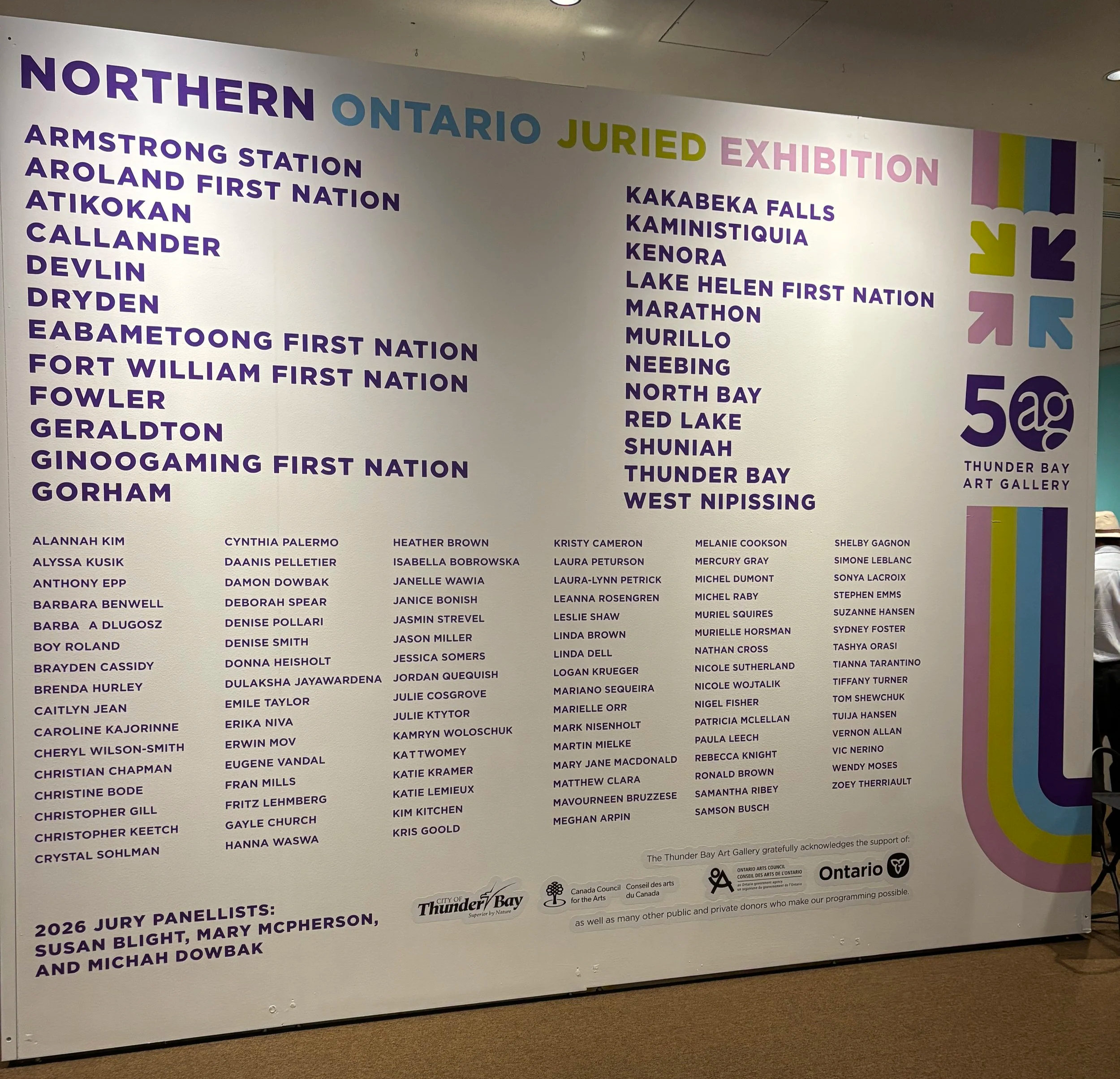 A large wall display at the Thunder Bay Art Gallery featuring lists of jury panelists and other acknowledgments, with colorful logos and headings.