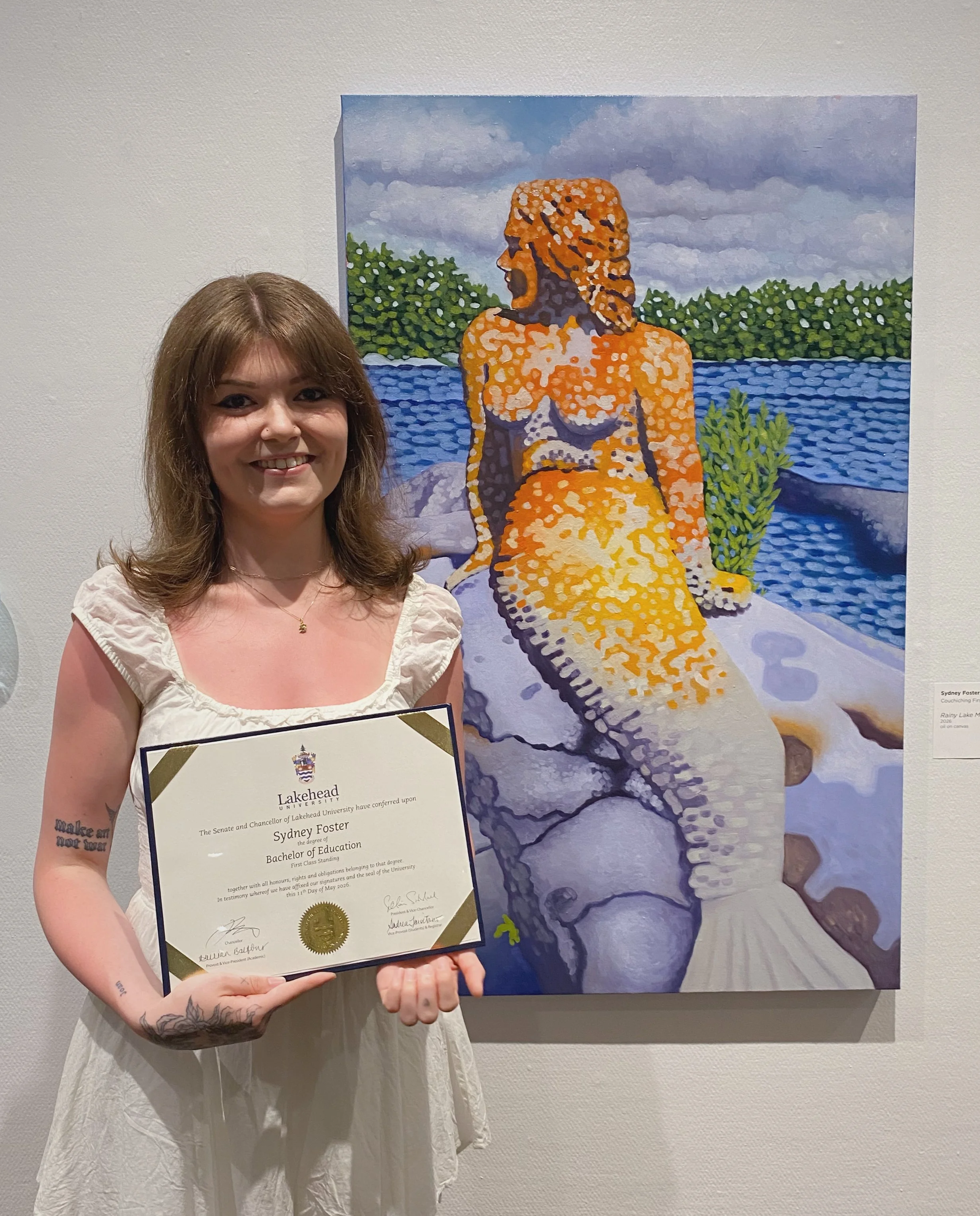 A young woman with tattoos holding a diploma while standing in front of a colorful abstract painting of a woman by a body of water with greenery and cloudy sky in the background.