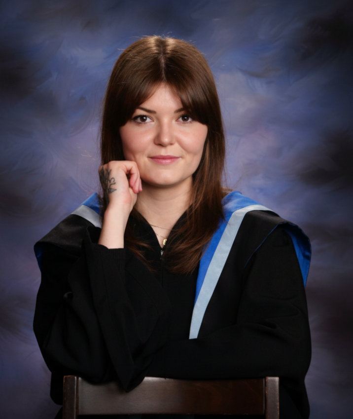 Woman in black graduation gown with blue and gray hood, seated against a dark, cloudy backdrop, smiling with her right hand resting on her chin.