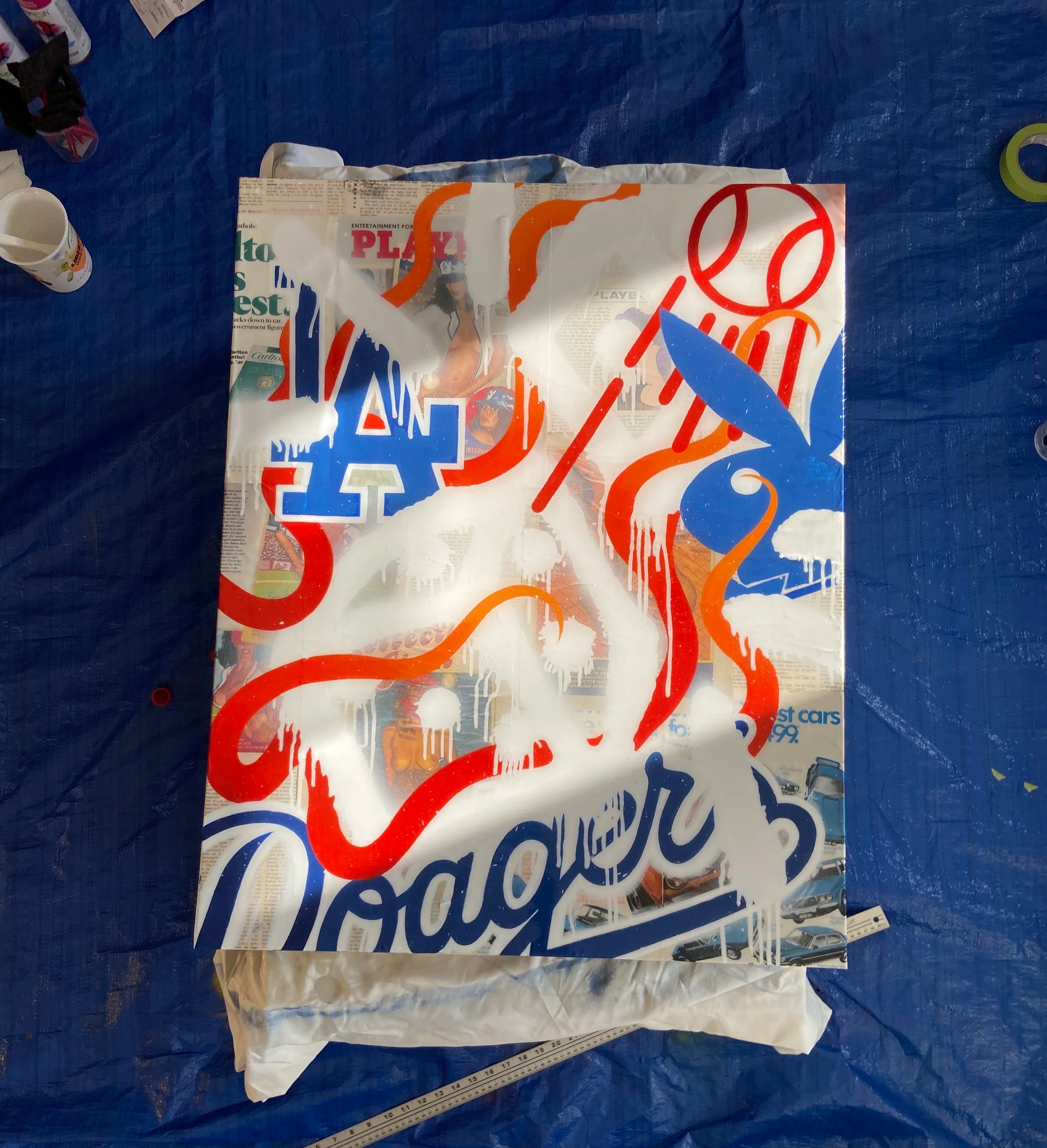 Colourful mixed media poster made of vintage magazines, with partial text and images of a woman, cars, and bold graphics, placed on a blue surface with a ruler and miscellaneous items nearby.
