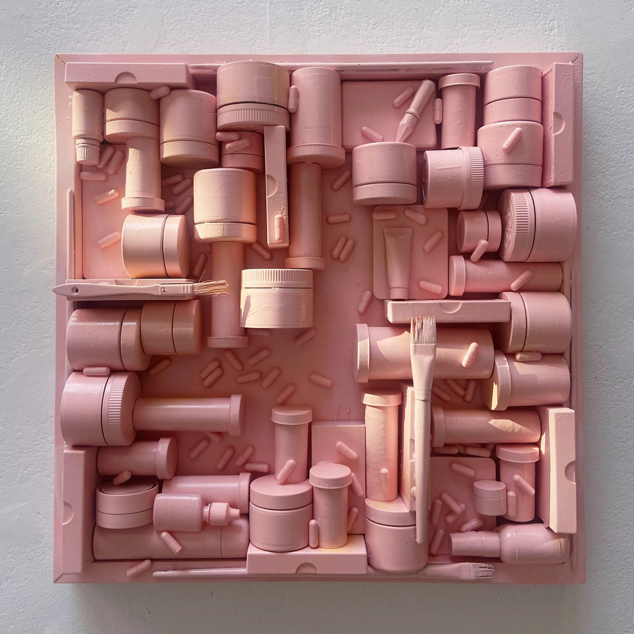 Pink art installation featuring various cylindrical objects, capsules, and small shapes arranged in a grid on a pink background.