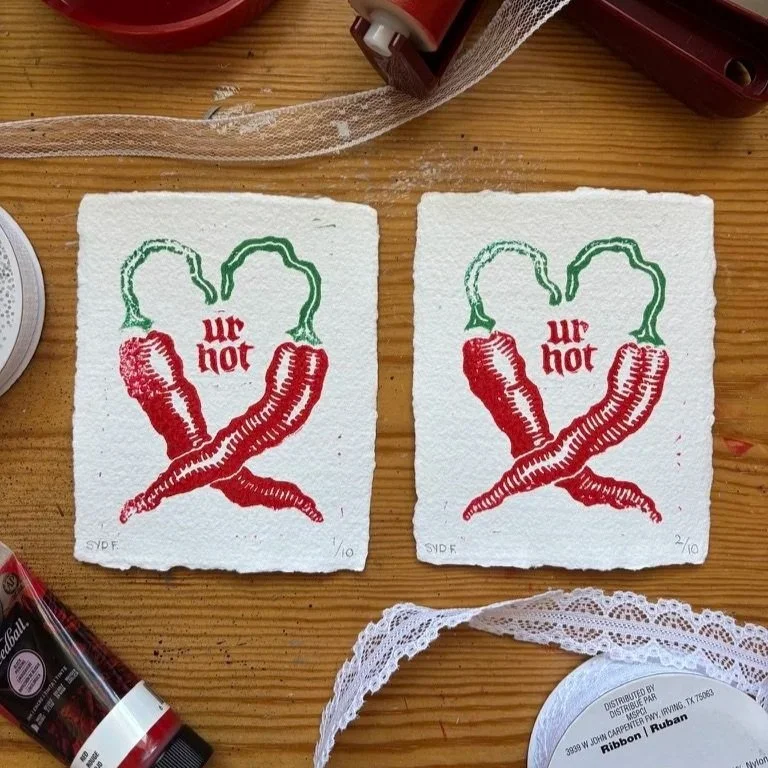 Two small pieces of paper with red and green chili peppers and the words "ur not" in red in the middle, placed on a wooden surface. Surroundings include a lace cloth and other craft supplies.