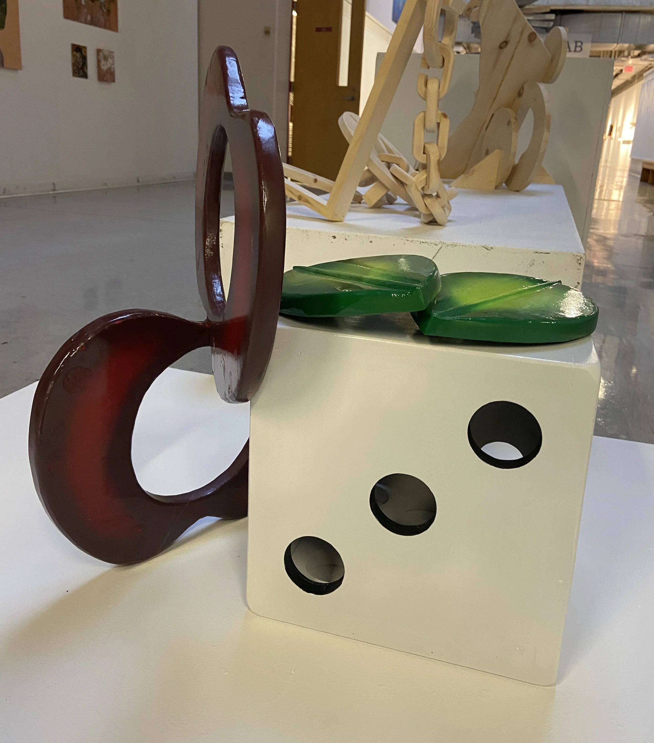 Close-up of a decorative display with a red, painted wooden bunny, green ceramic leaves, and a white block with black circular cutouts. In the background, part of a wooden sculpture and gallery wall are visible.