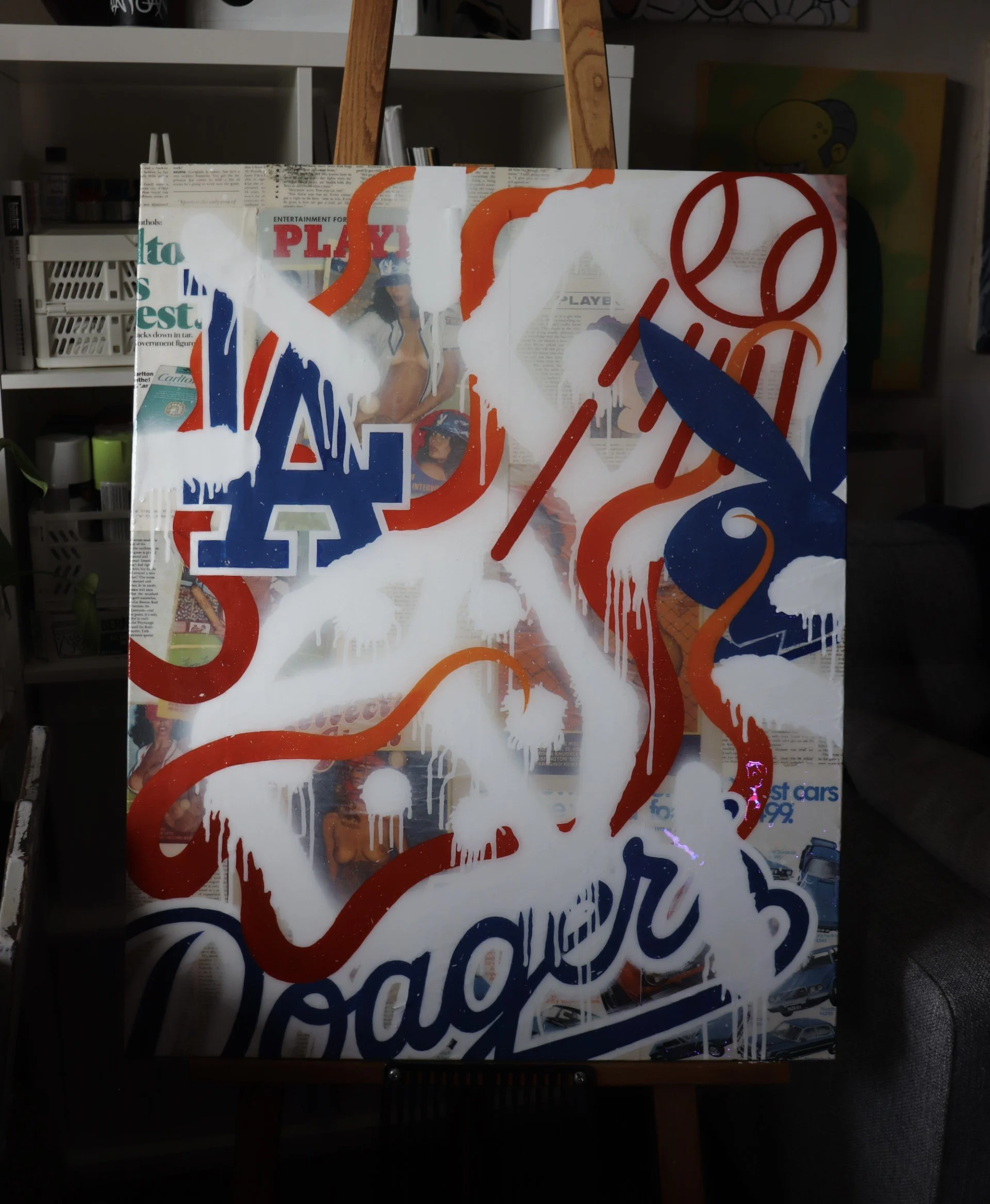 A glass panel with red, white, and blue graffiti-style designs covering newspaper clippings behind it, displayed on an easel in a room with shelves and artwork.