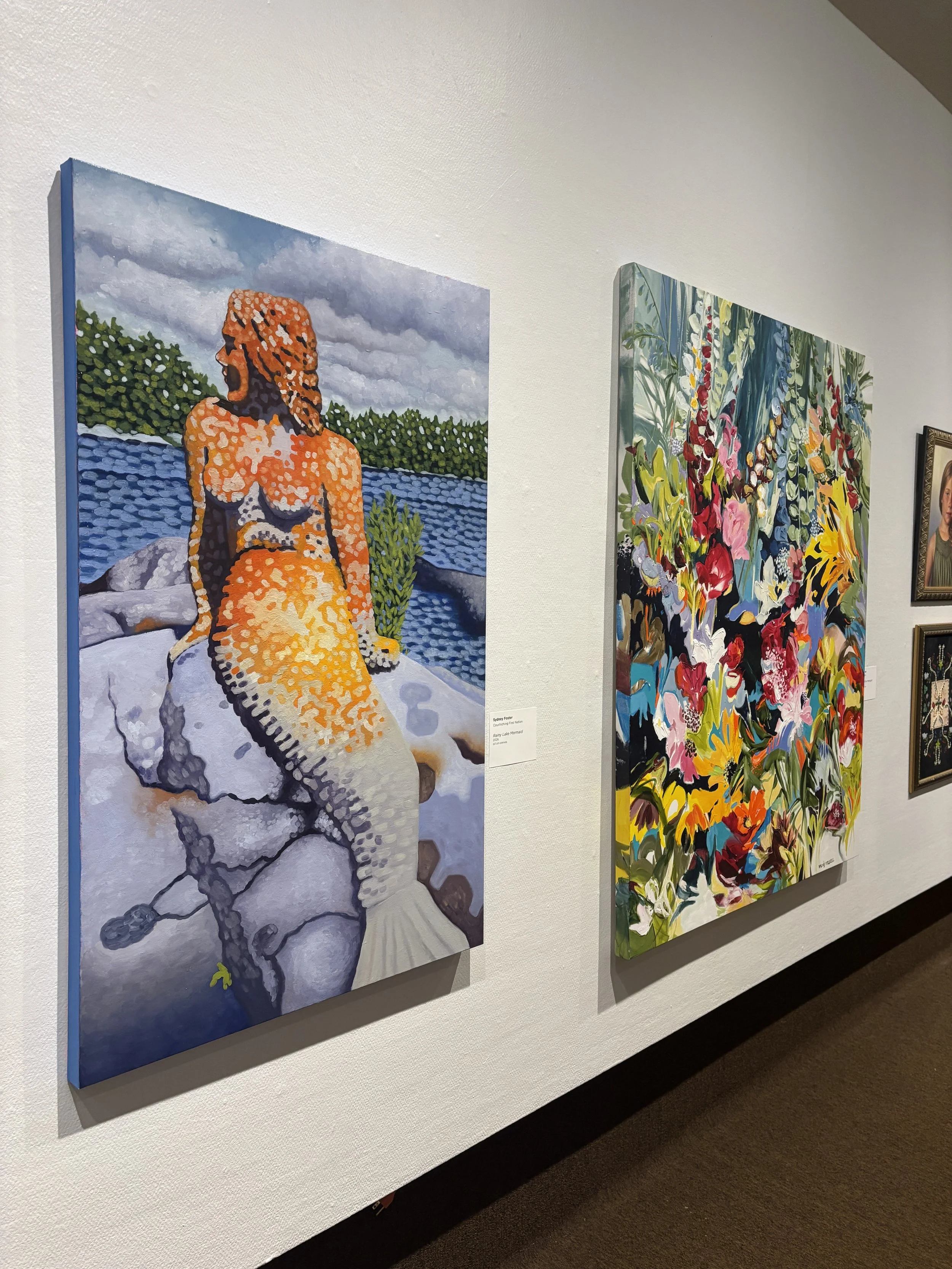 Two colorful paintings on a gallery wall. The left painting depicts a woman with reddish hair sitting outdoors by a body of water with cloudy skies in the background. The right painting features an abstract floral design with vibrant colors.