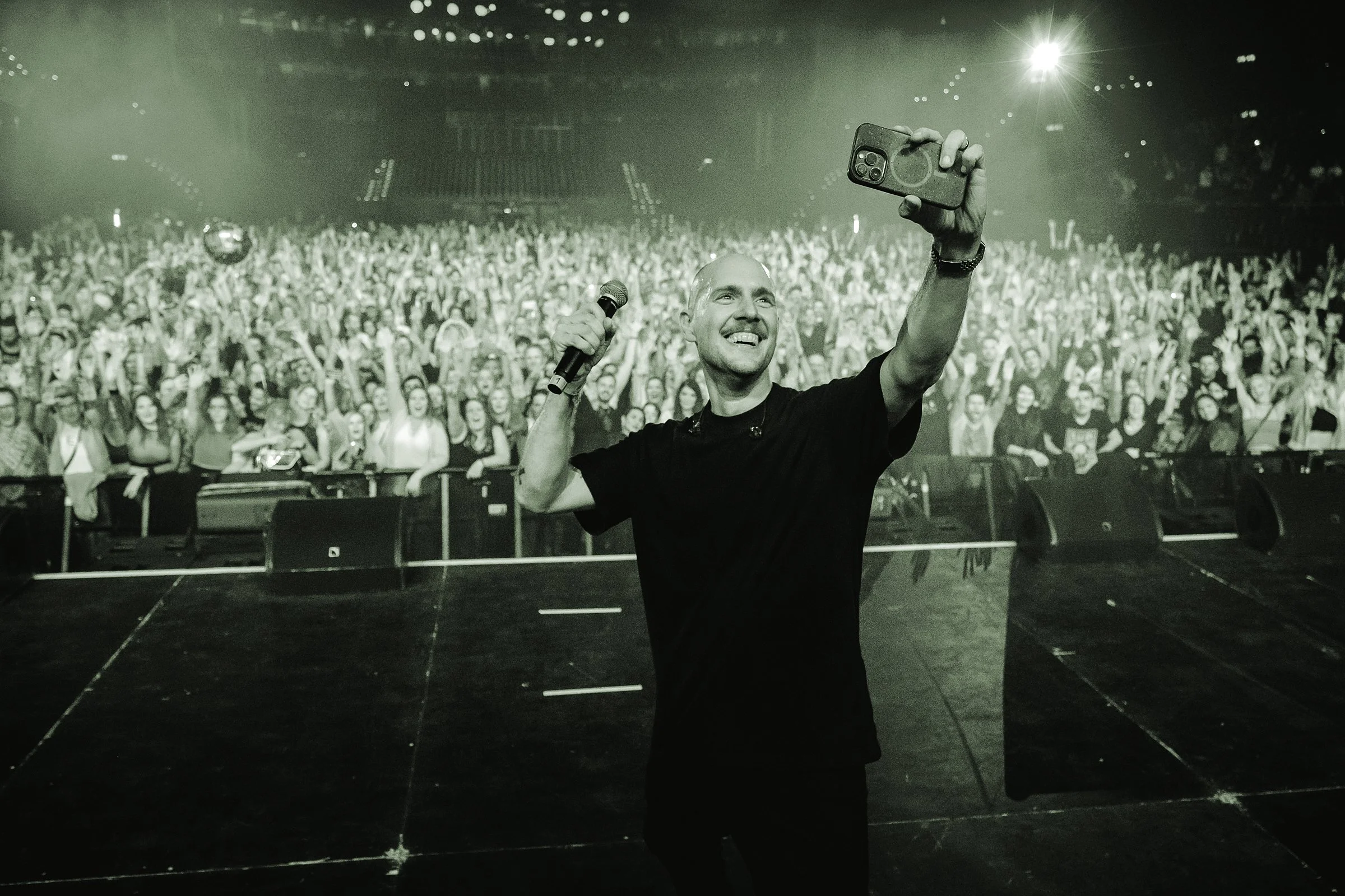 Ein Mann auf einer Bühne bei einem Konzert, der in die Kamera selfie macht, während die Menge im Hintergrund feiert.