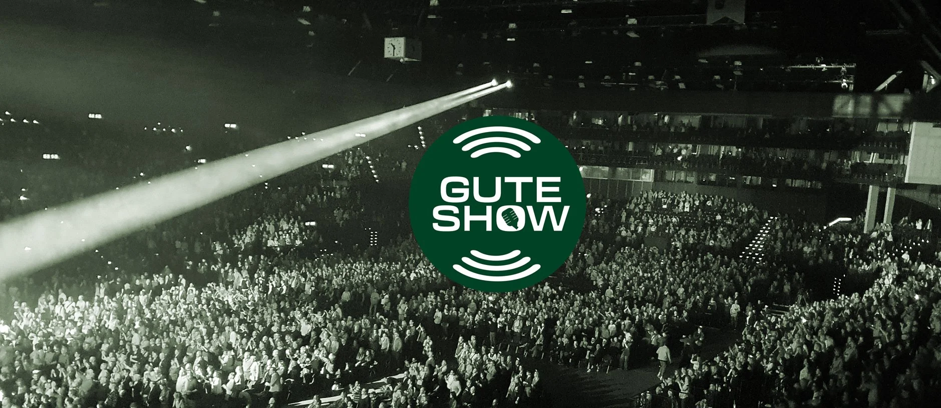 Ein ausverkauftes Konzertstadion mit einem großen Publikum, mit einem grünen Rundlogo in der Mitte, das die schwarzen Worte 'GUTE SHOW' und eine Mikrofontasche enthält, umgeben von weißen Signalen, die eine Klangwellen-Darstellung darstellen.