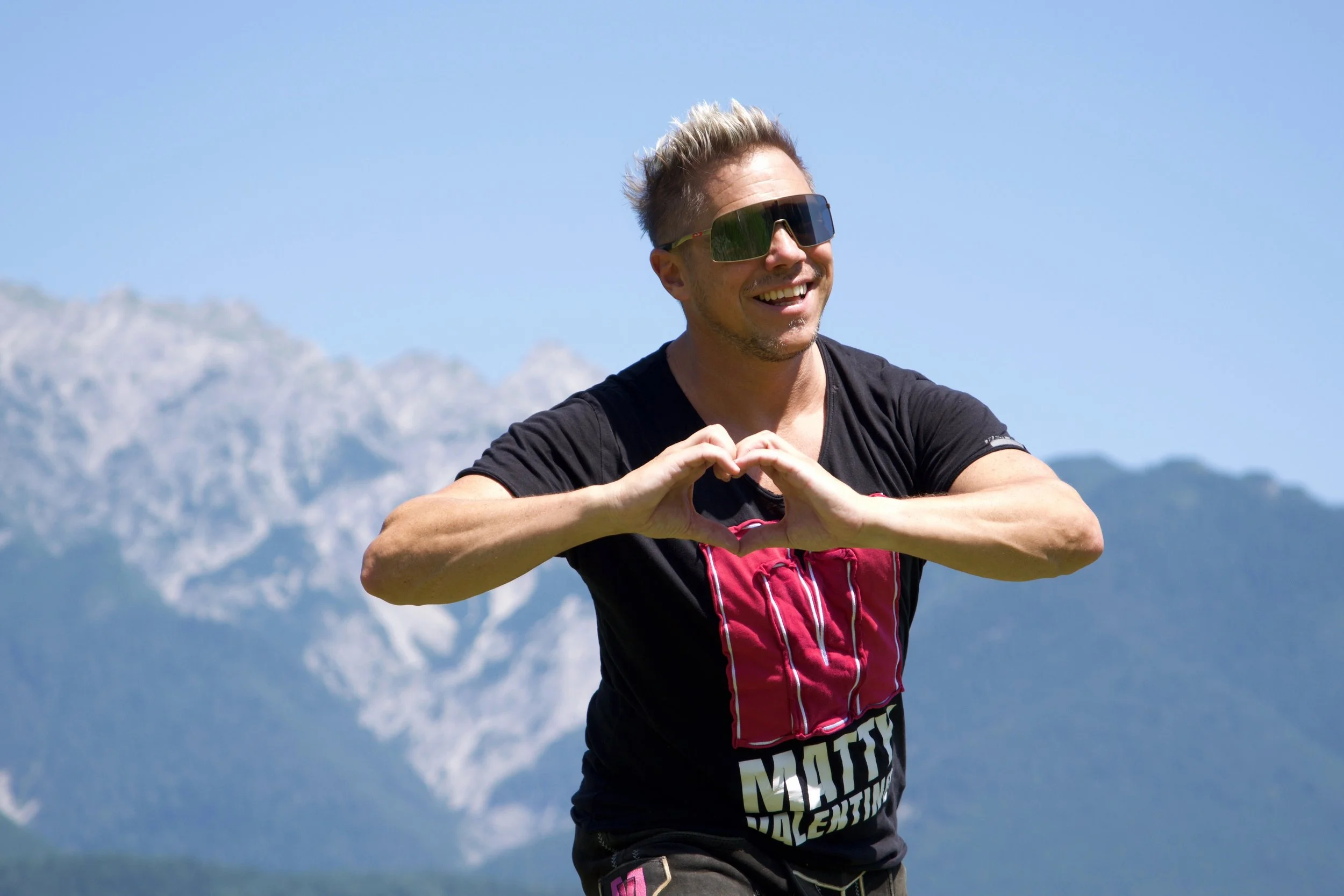 Junge Mann macht Herzform mit den Händen vor Bergen und blauen Himmel.