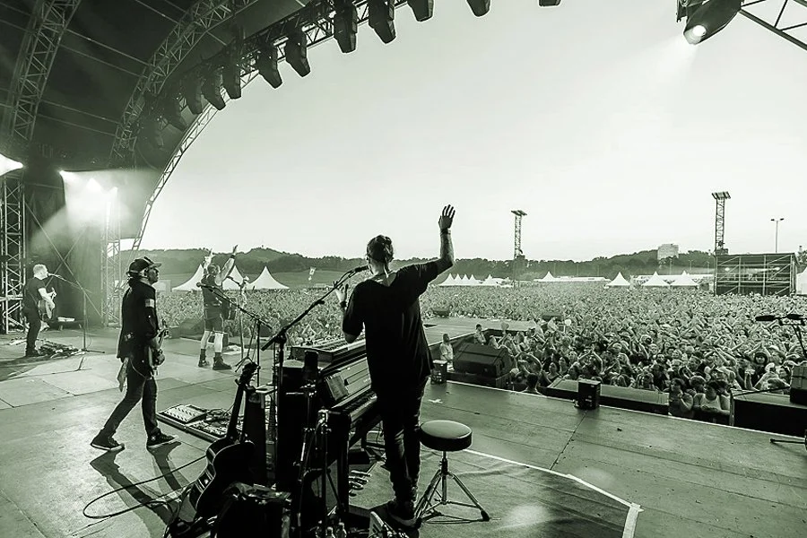 Musiker auf einer Bühne während eines Open-Air-Konzerts, das Publikum steht vor der Bühne auf einer weiten Wiese.