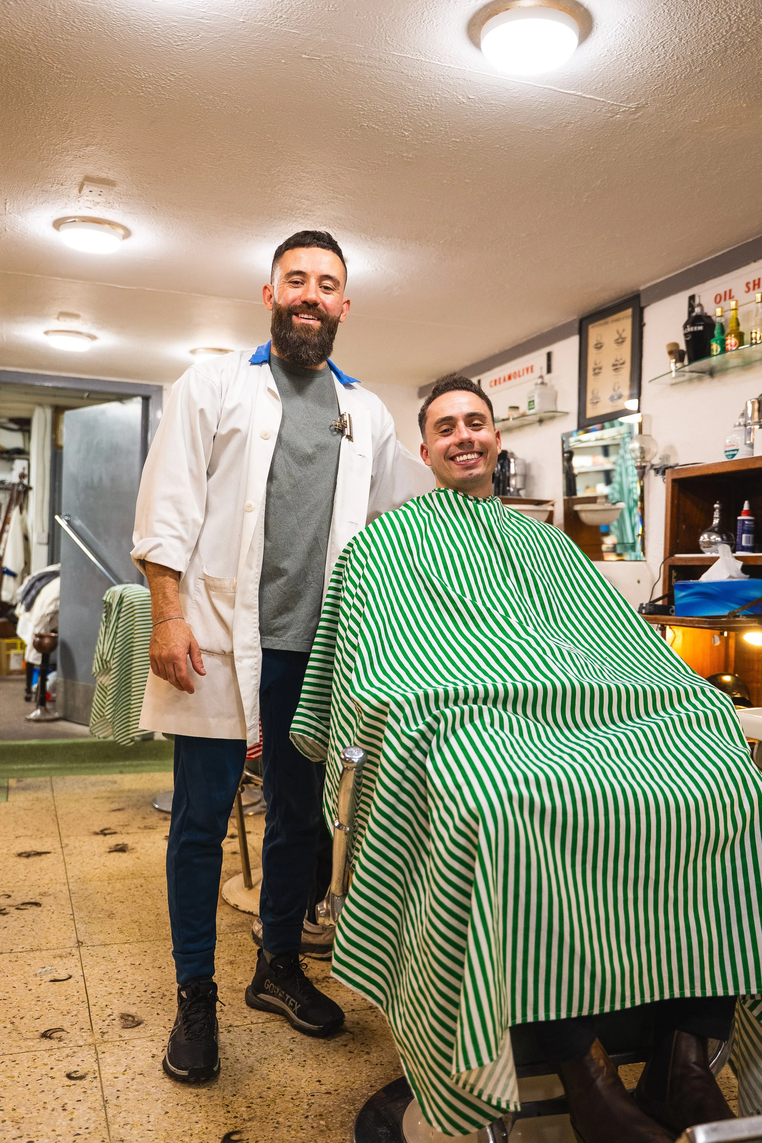 A man with a beard getting a haircut in a barbershop, smiling and sitting in a barber chair with a green and white striped cape, while the barber, standing next to him, is wearing a white coat and smiling.