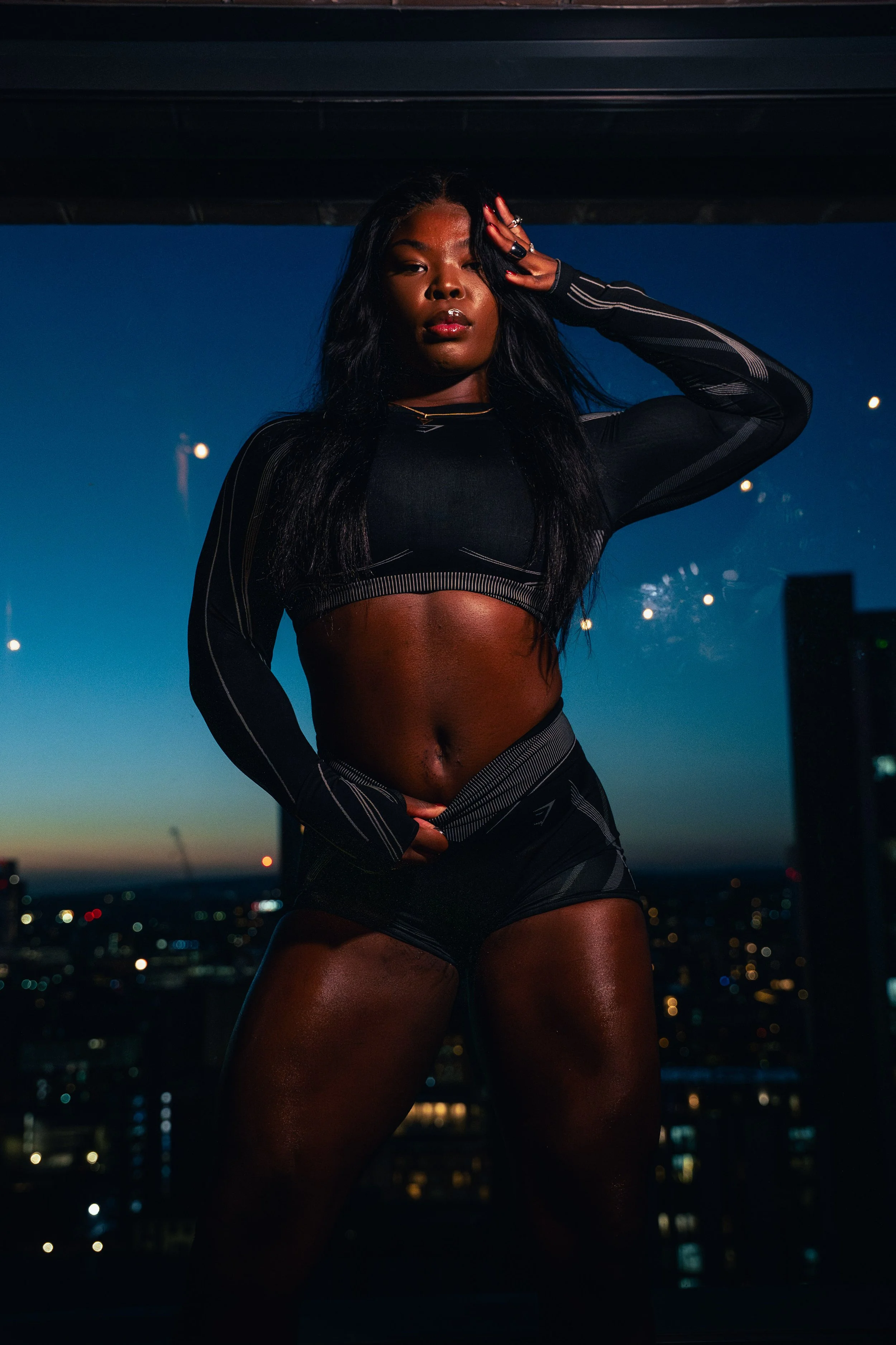 A woman with dark skin and long black hair standing indoors at night, with city lights visible through the window behind her. She is wearing a black sports bra and matching shorts, and posing with her hand on her head.