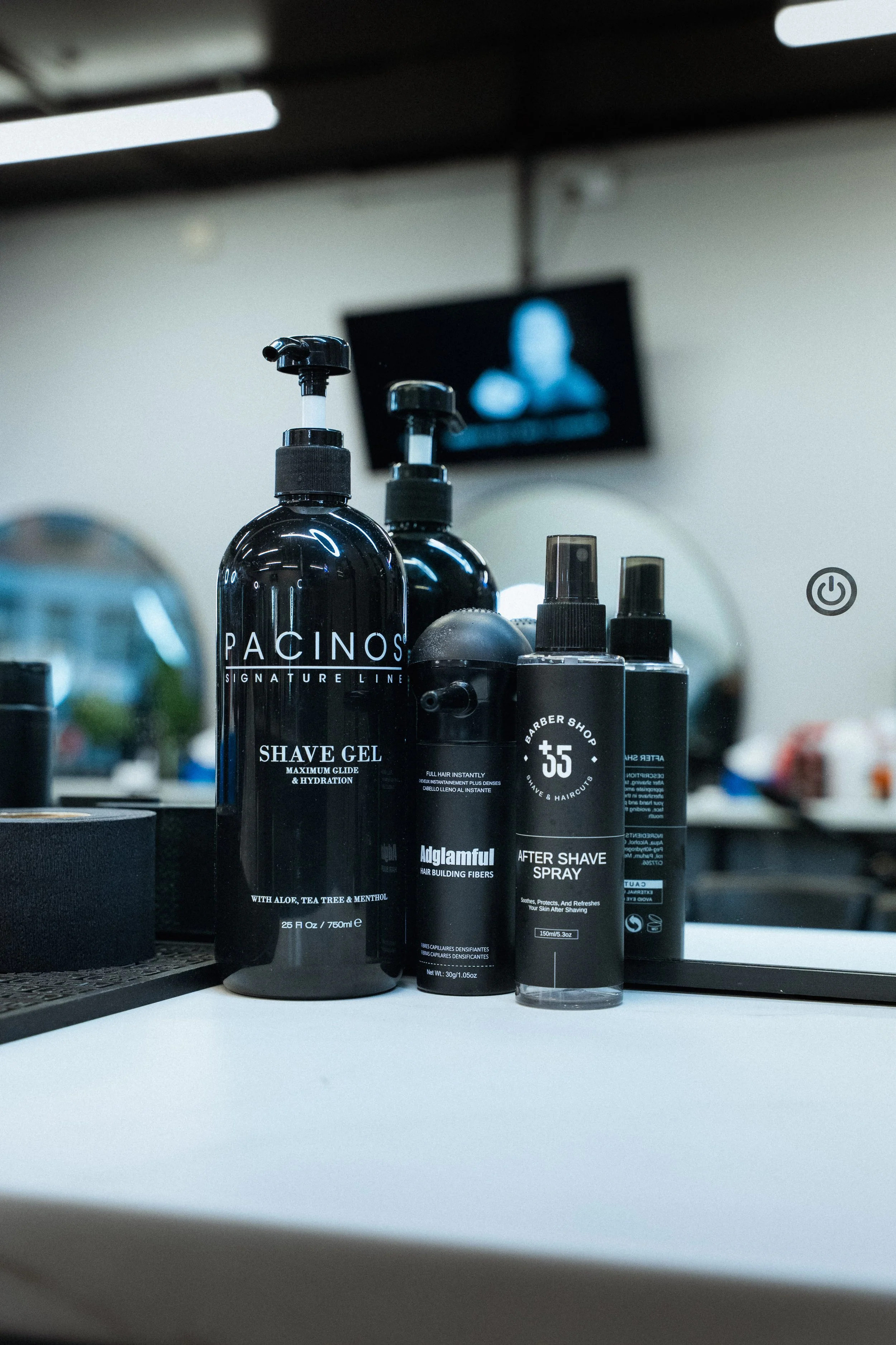 A collection of men's grooming products on a barbershop counter, including shave gels, after shave spray, and hair building fibers, with a mirror and a television screen in the background.