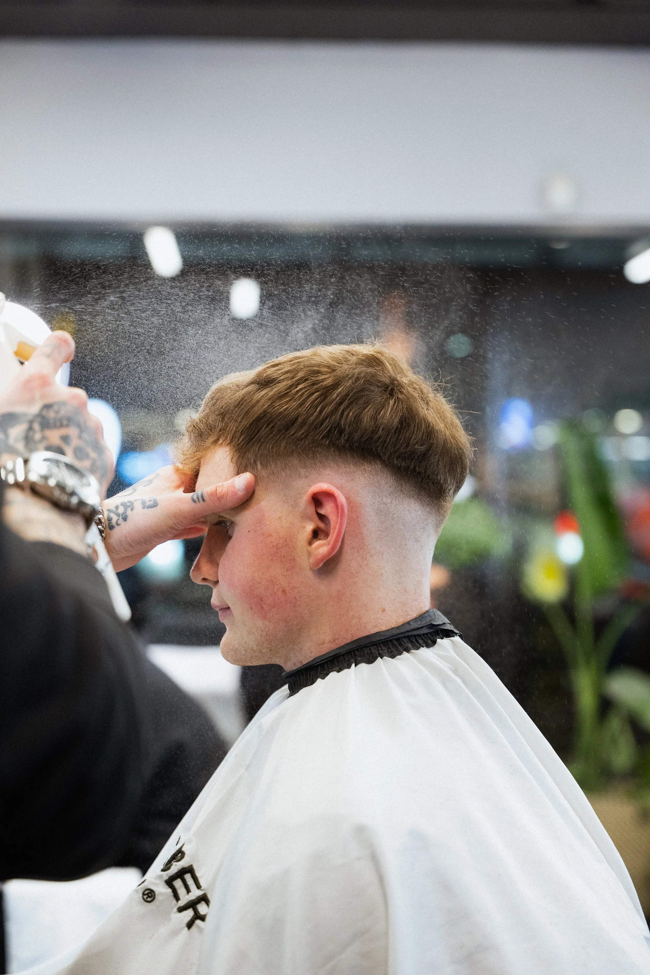 A person getting a haircut at a barbershop, with the barber spraying water on their hair. The individual has short brown hair and is wearing a white barber cape.