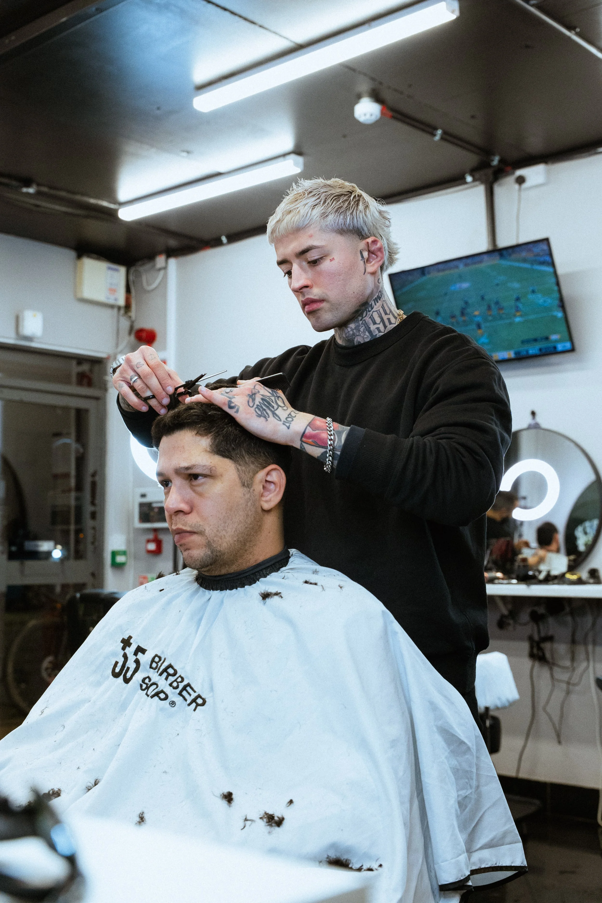 Barber cutting a man's hair in a salon, with a television screen showing a sports game in the background.