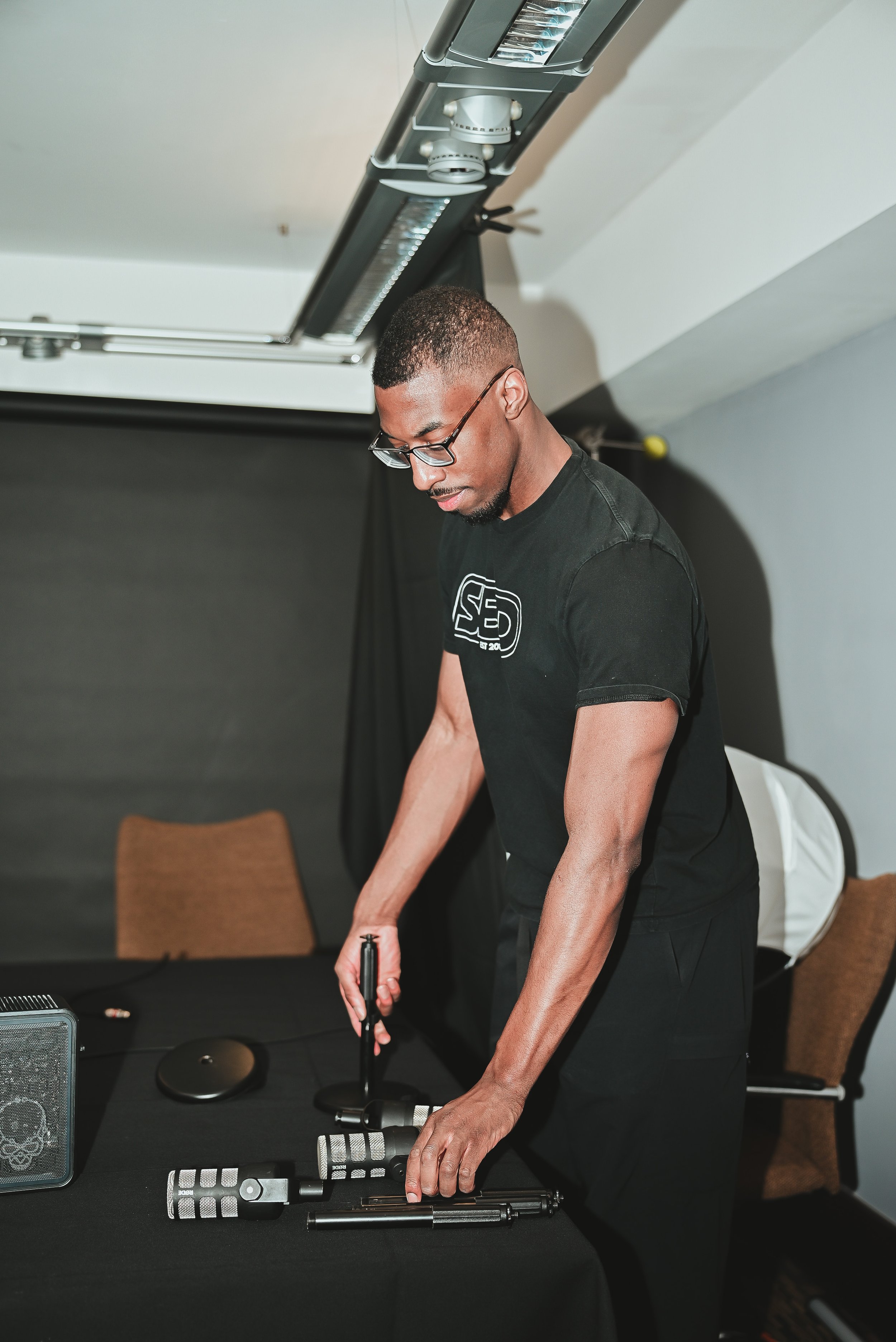 A man with glasses and a black t-shirt is standing at a table, handling equipment including a microphone and various audio devices in a room with several chairs and a black backdrop.