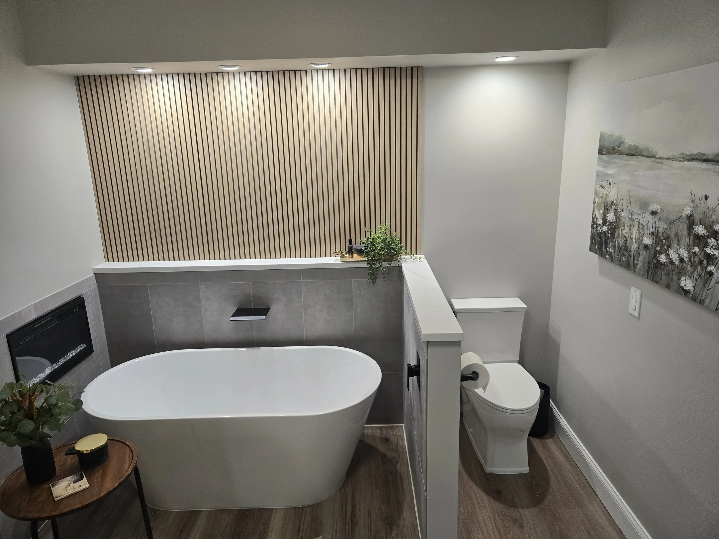 Modern bathroom with a freestanding oval bathtub, a toilet, and a wooden accent wall with vertical slats. There is a small table with decor and a plant, and a large painting on the wall.
