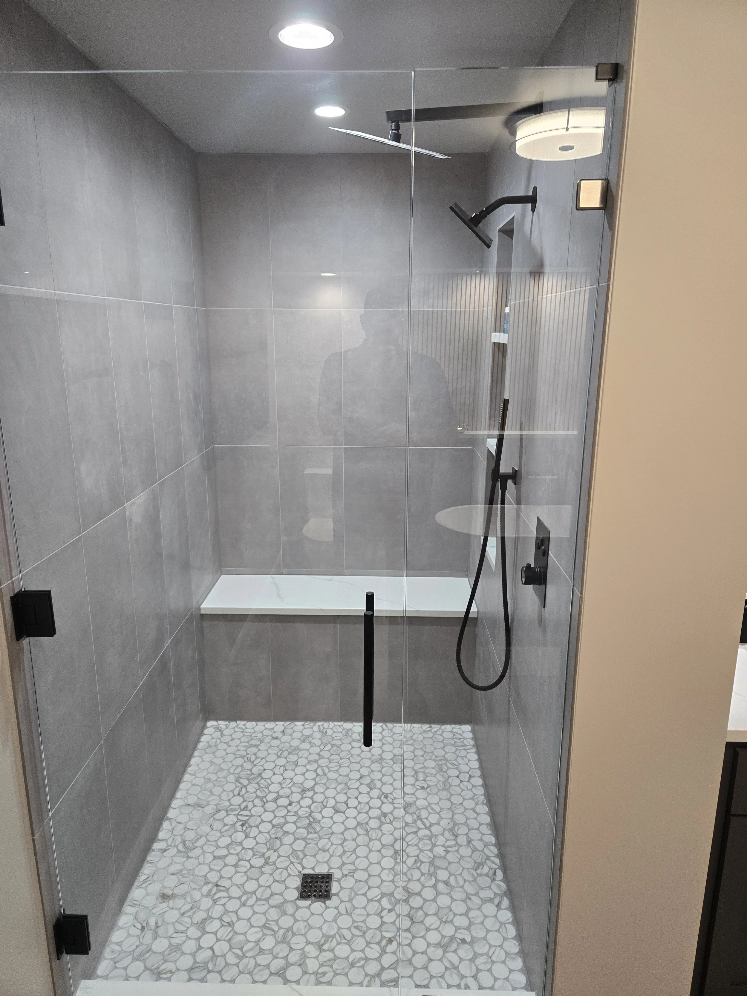 Modern glass shower enclosure with gray tiles, pebble-patterned shower floor, black shower fixtures, and a ceiling light in a bathroom.